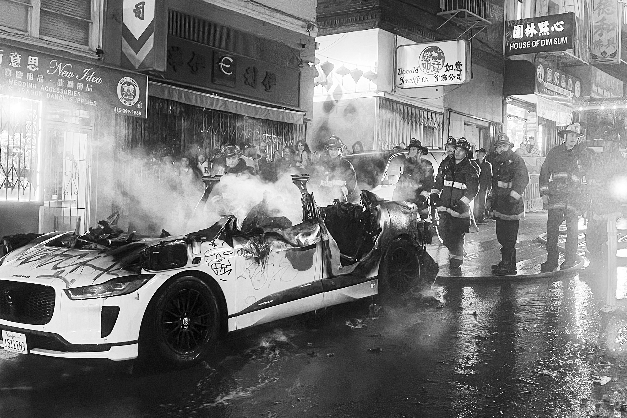 A black and white photo of a burned-out Waymo autonomous car surrounded by firefighters amidst steam on a city street at night.