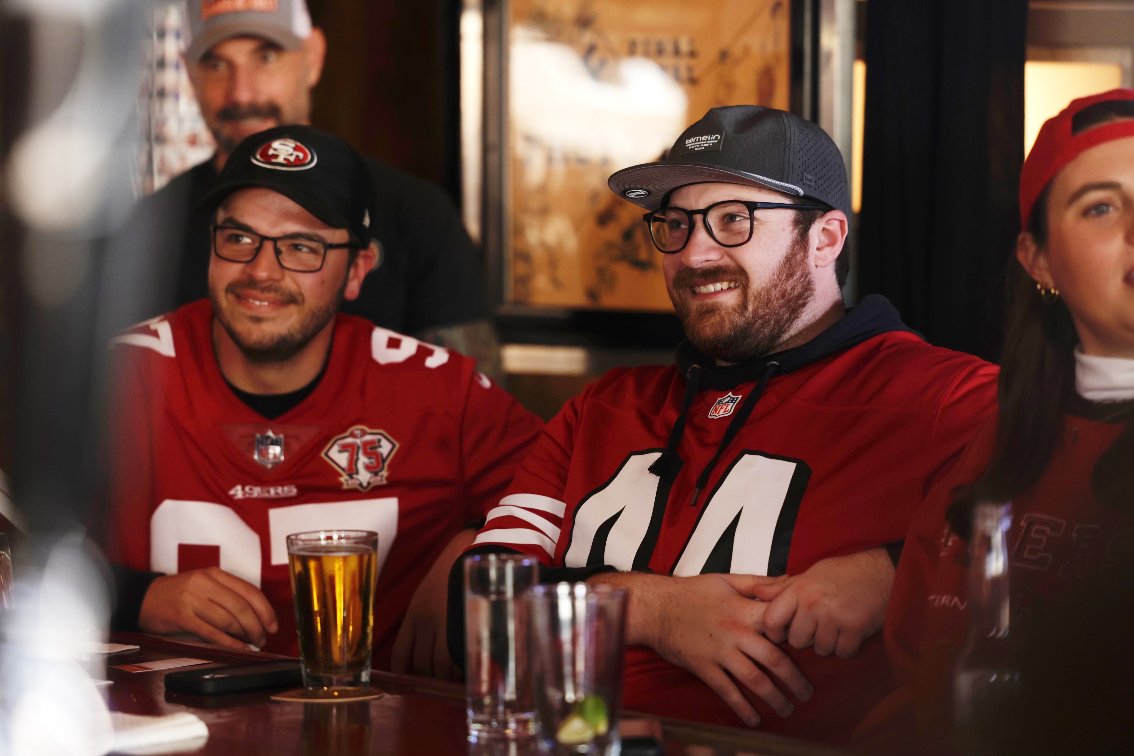 Fans in a bar wear 49ers jerseys and caps, smiling, with drinks on the table.