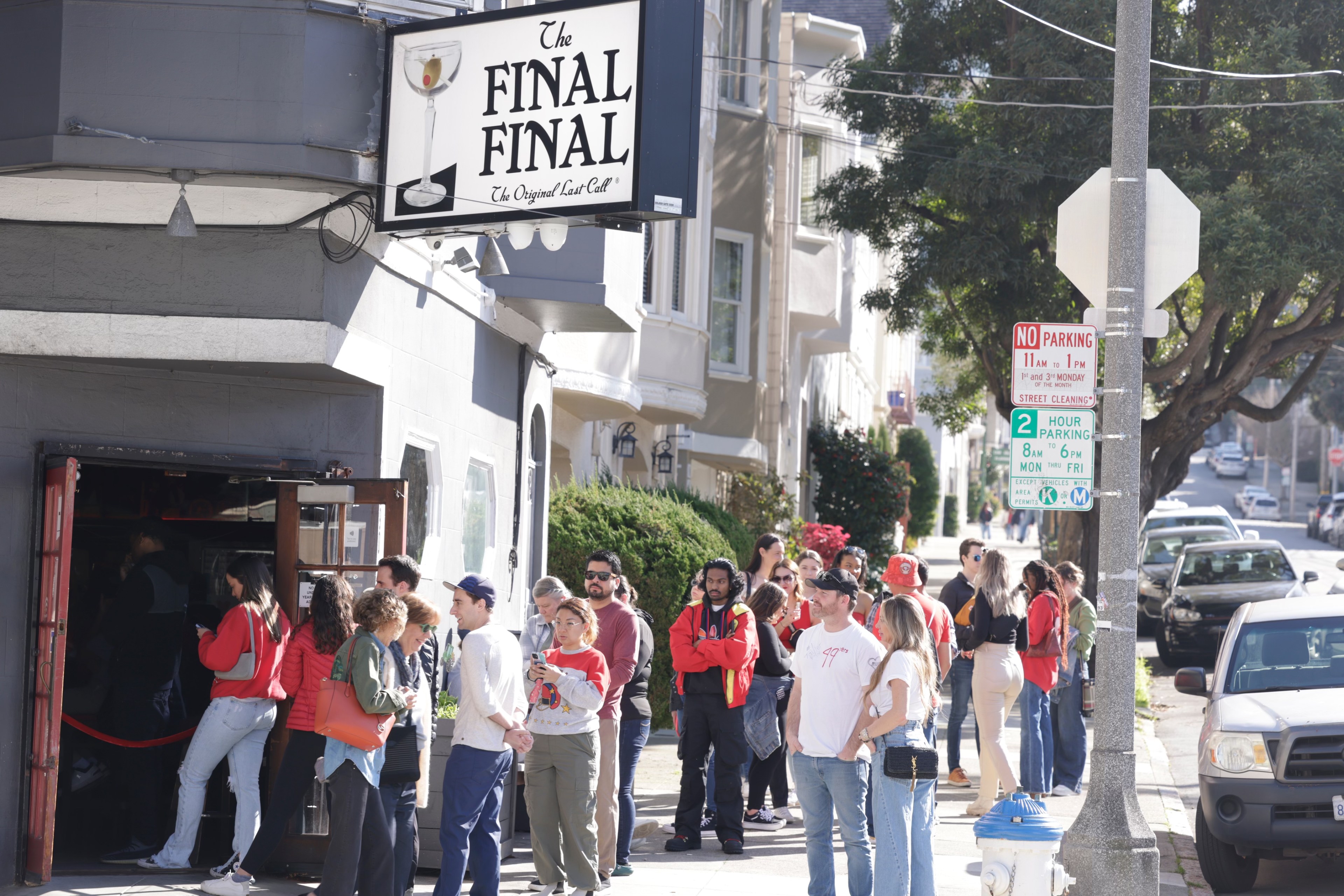 A bustling line of people waits outside &quot;The Final Final&quot; bar on a sunny street corner.