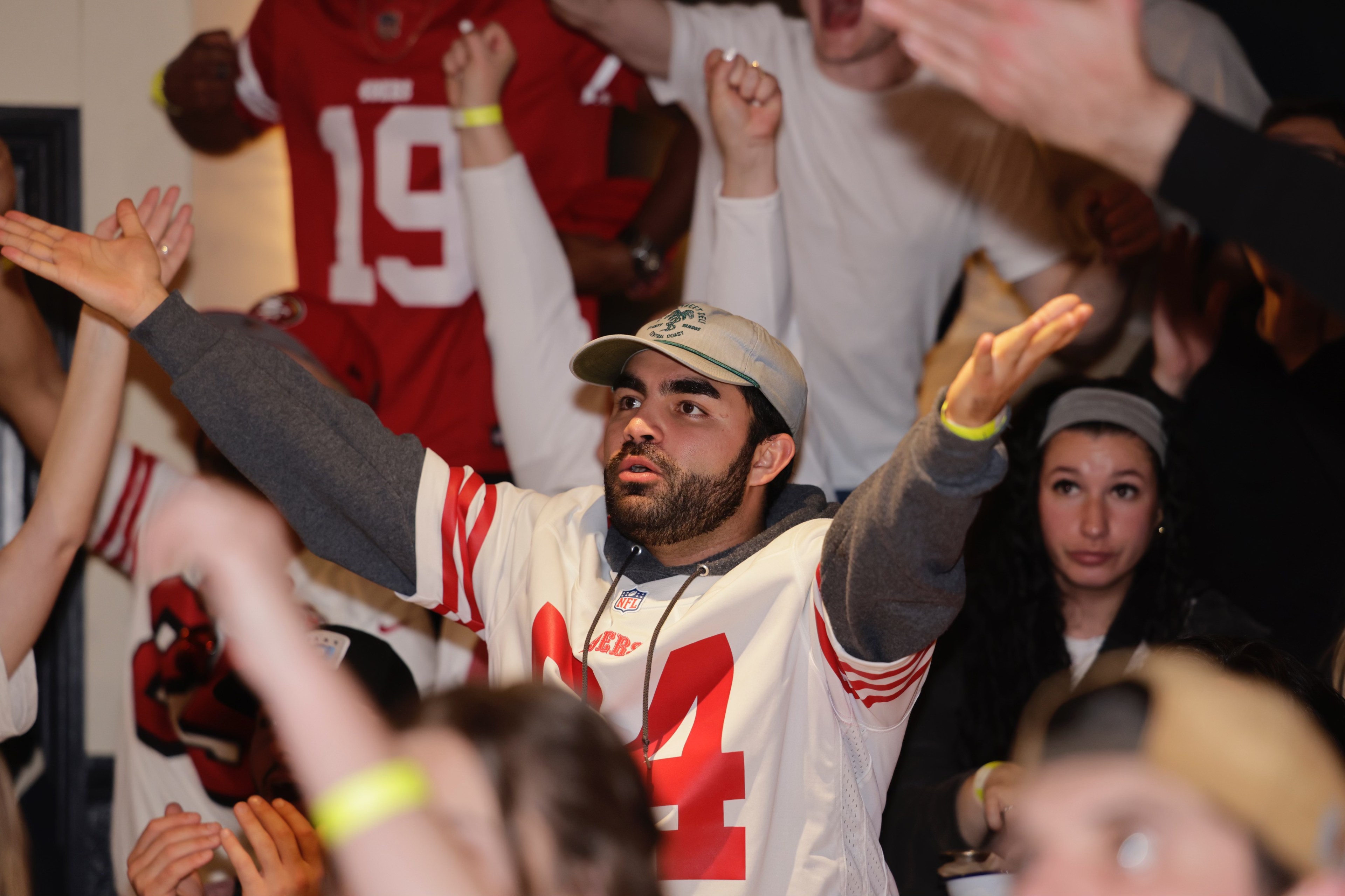 A crowd with raised hands; a man in a football jersey is in focus, signaling with serious expression.
