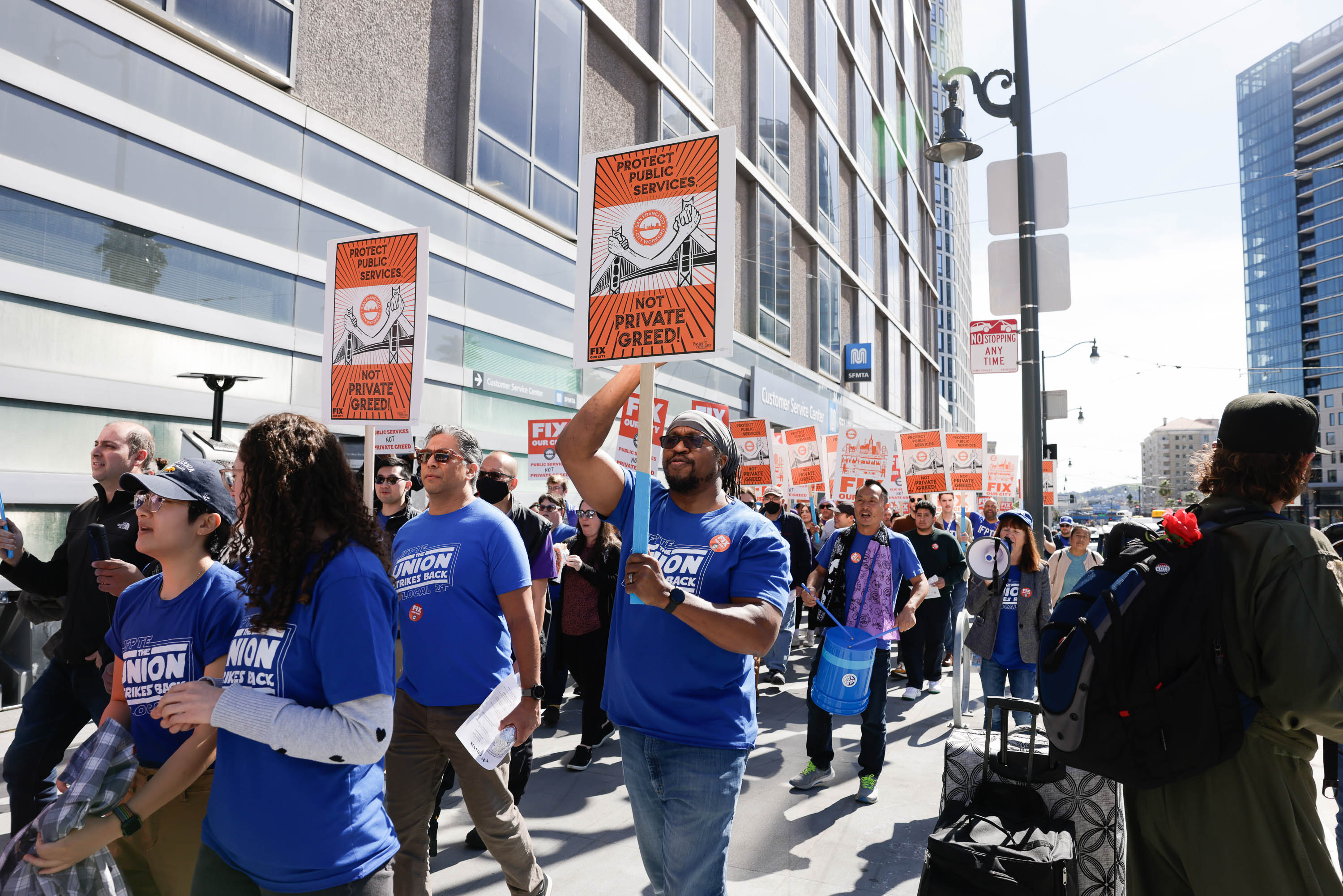 People marching in a protest with signs and blue union shirts, rallying against privatization.
