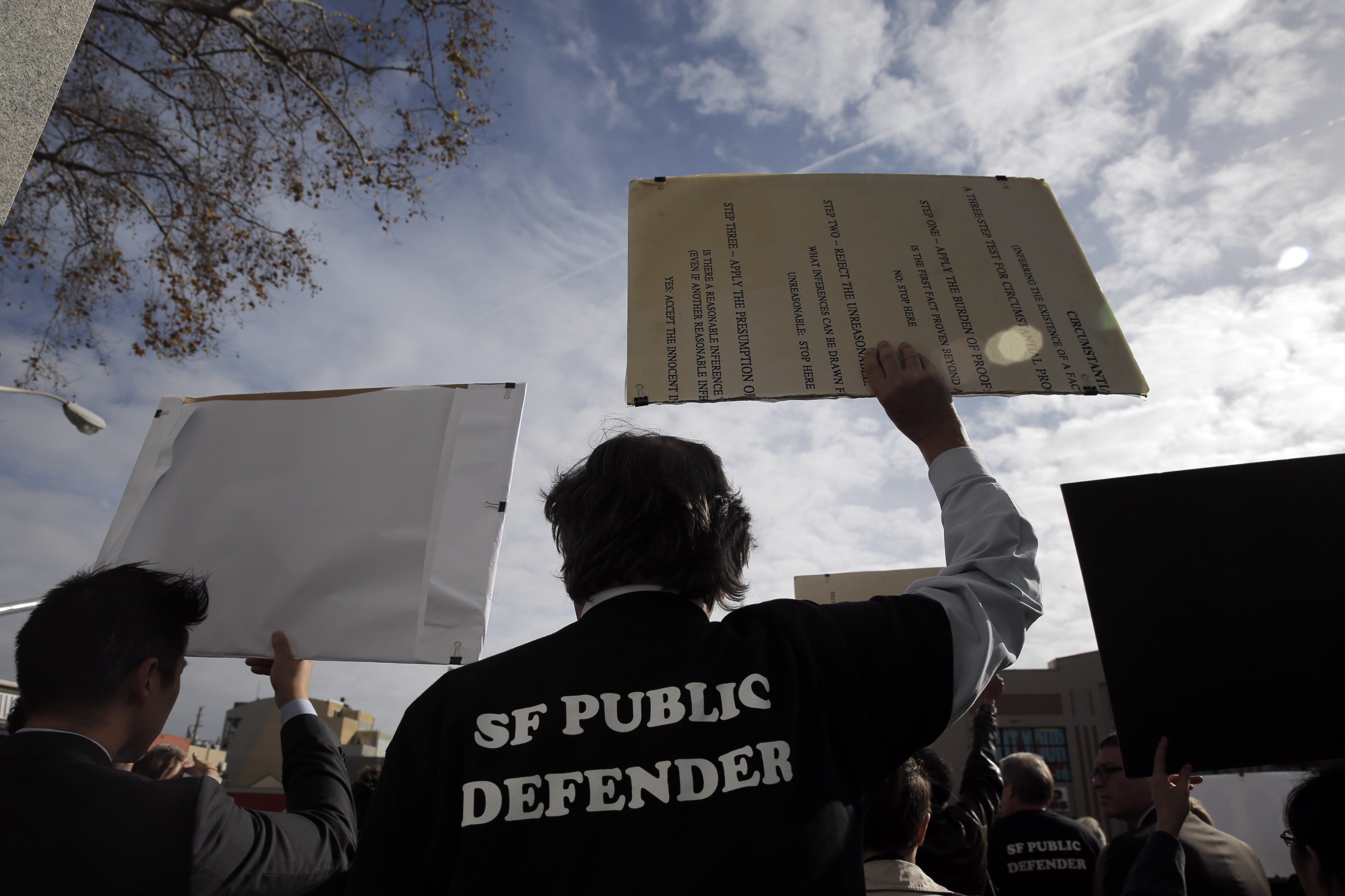 Person in a &quot;SF Public Defender&quot; jacket raises a sign at a protest under a cloudy sky.