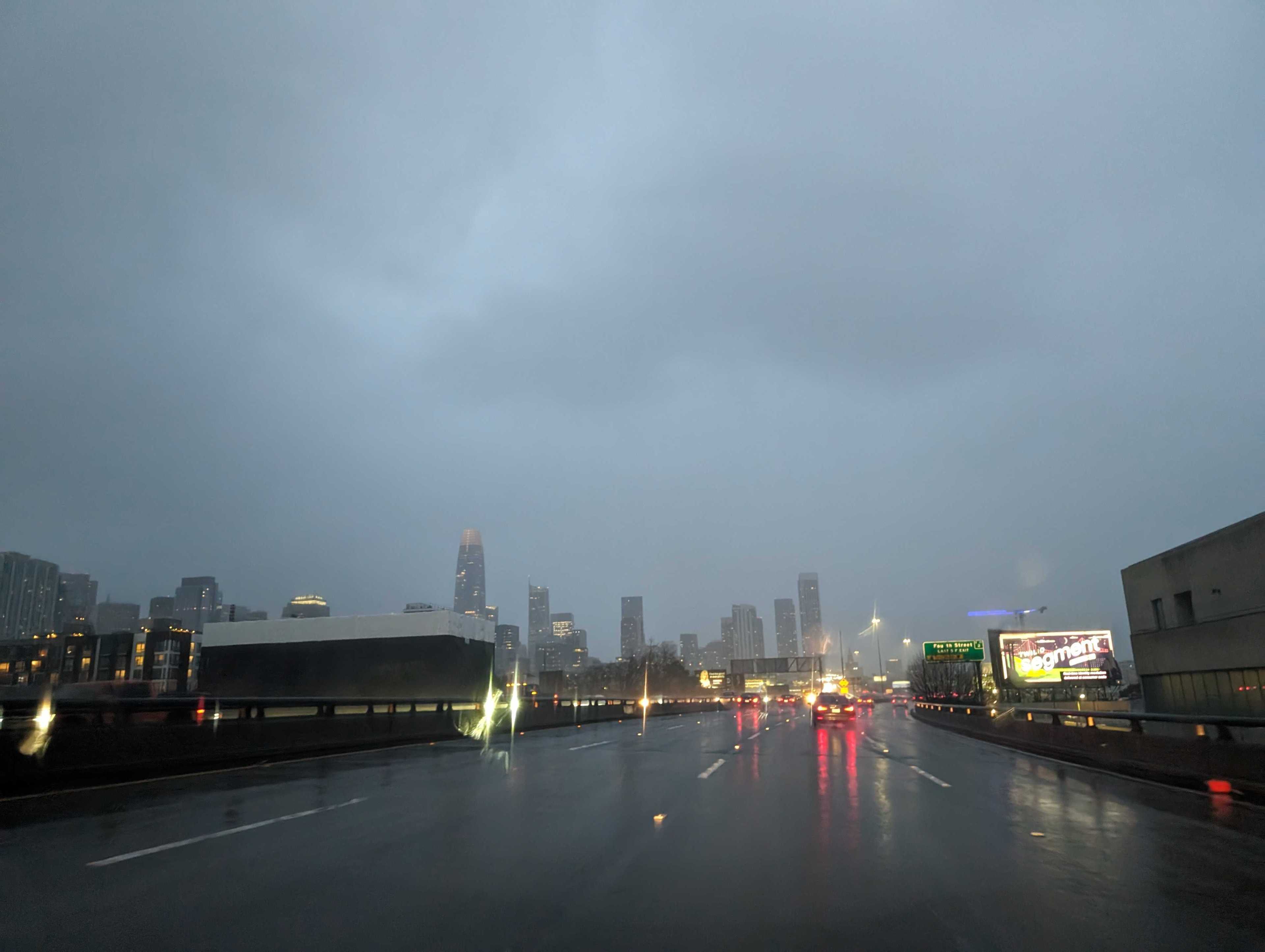 A city skyline nearby as seen from an elevated freeway with sparse traffic in rainy twilight.