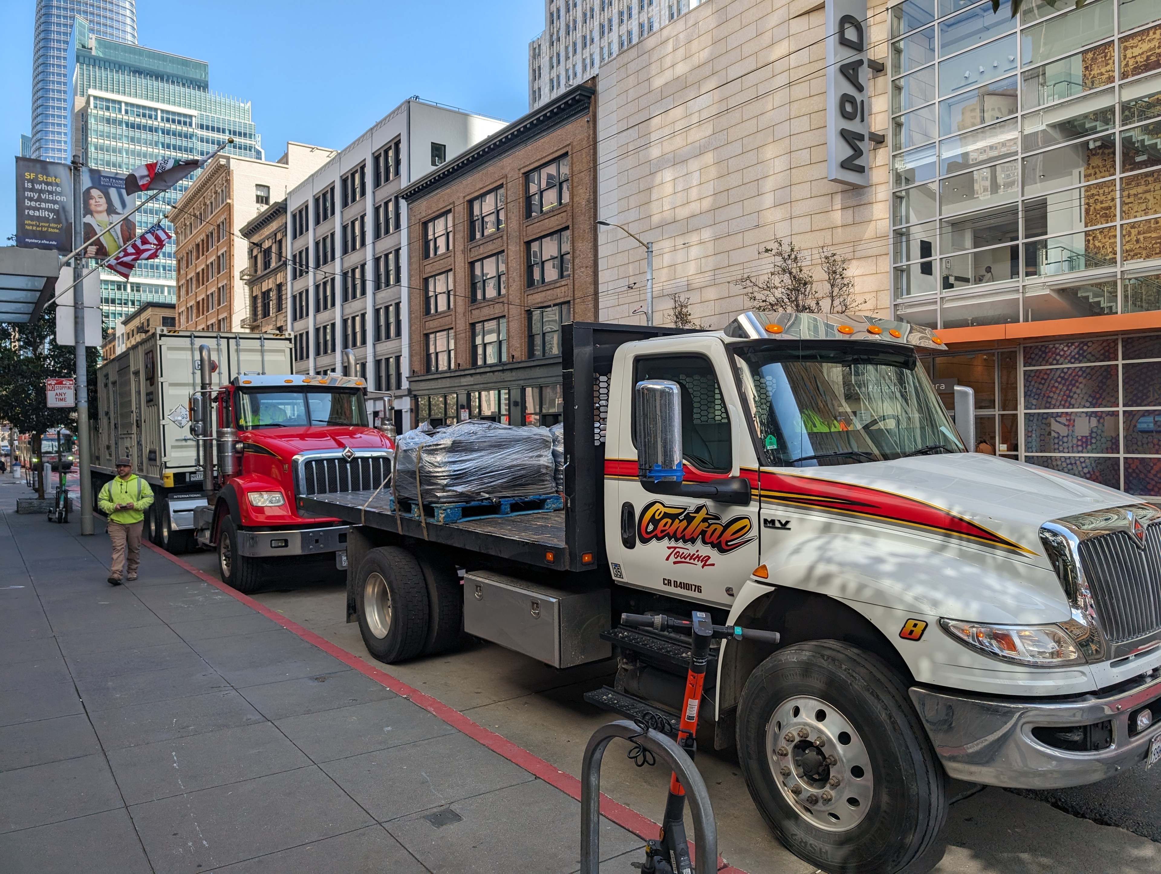 A big-rig truck and a towing truck bearing equipment park outside a city building at a red painted curb.