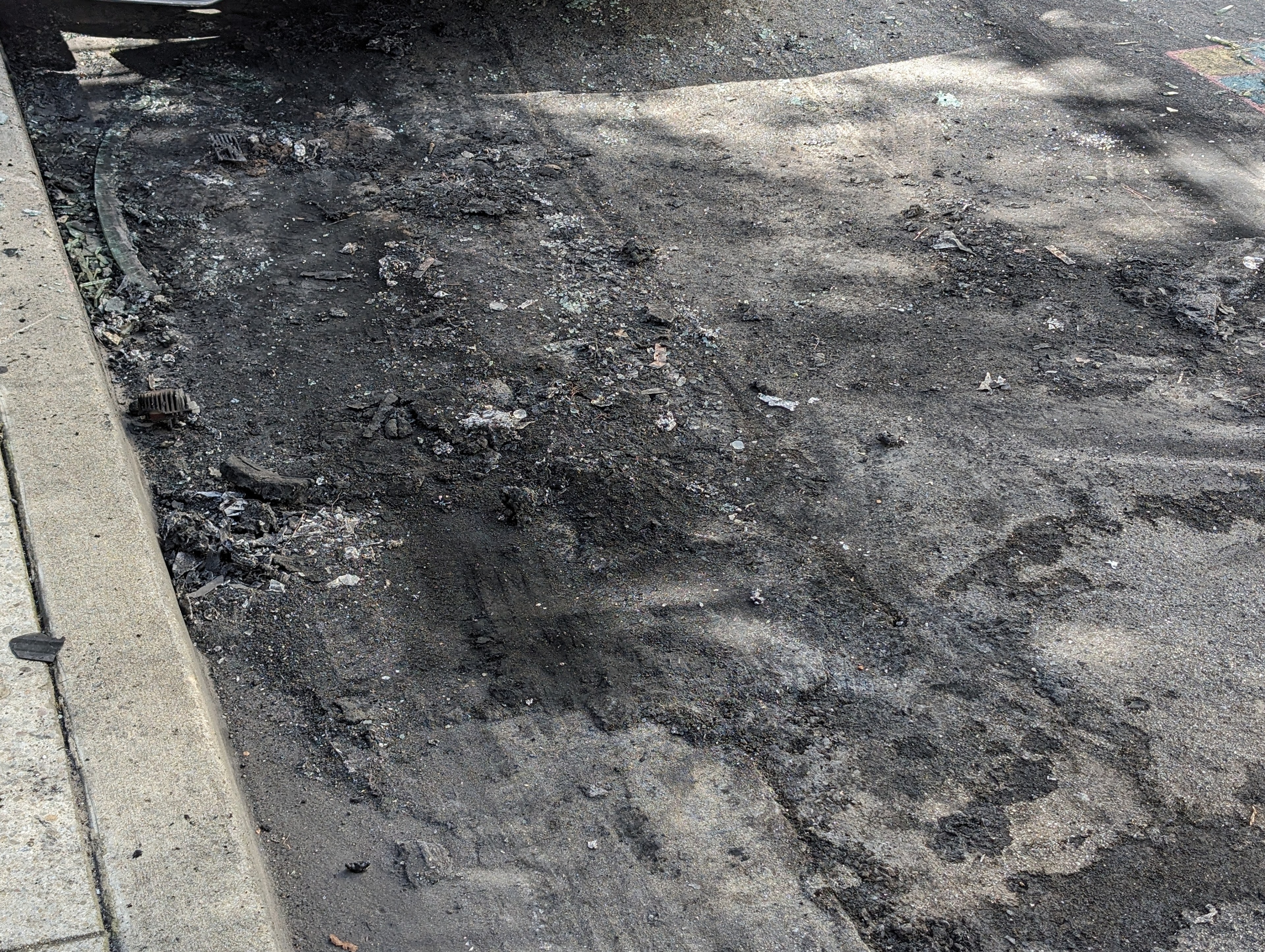 Charred ashes and vehicle parts remain in the roadway by a sidewalk curb.