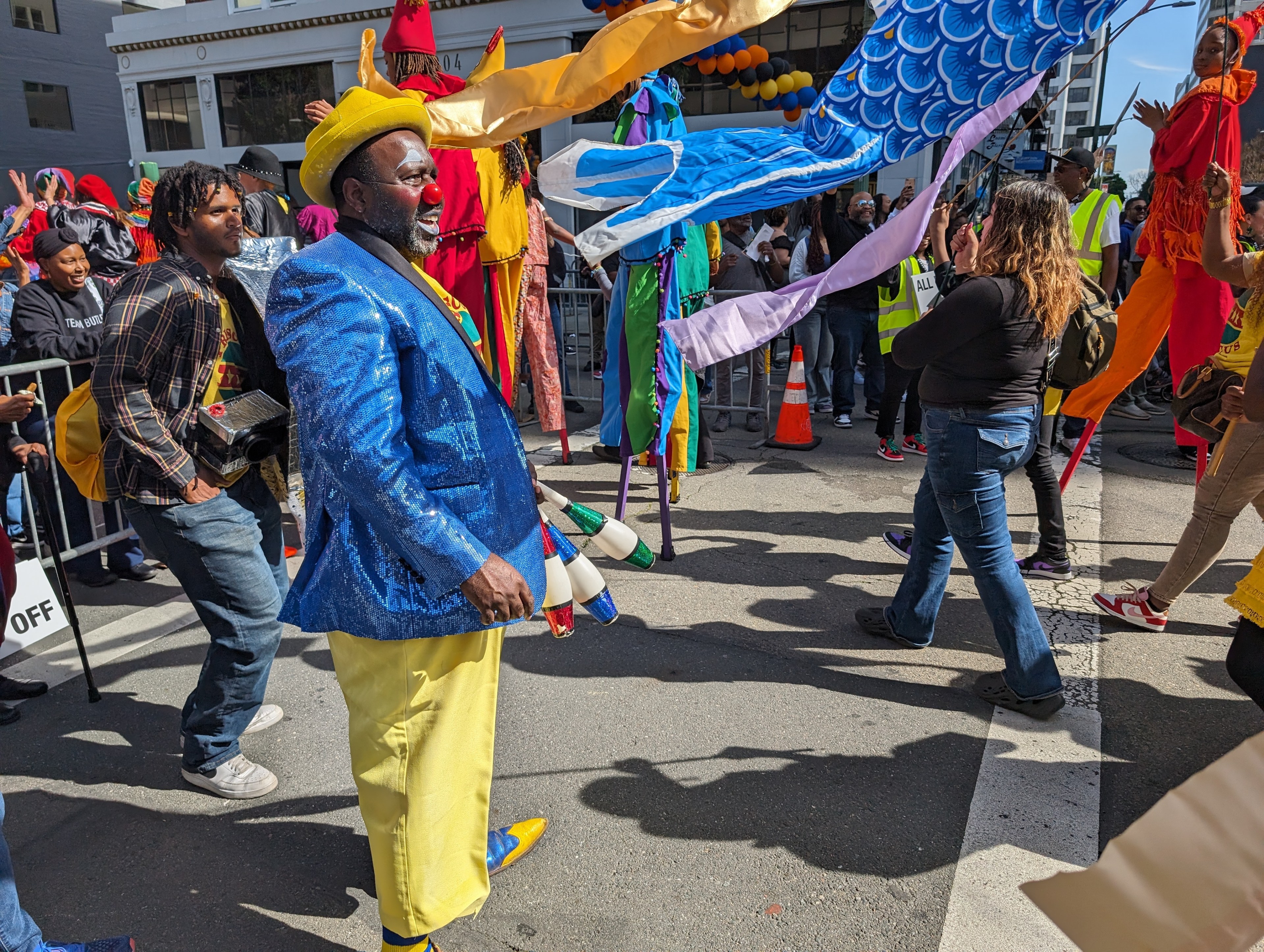 A man in brightly colored clothing and a red clown nose smiles as youngsters on stilts pass him in a parade.