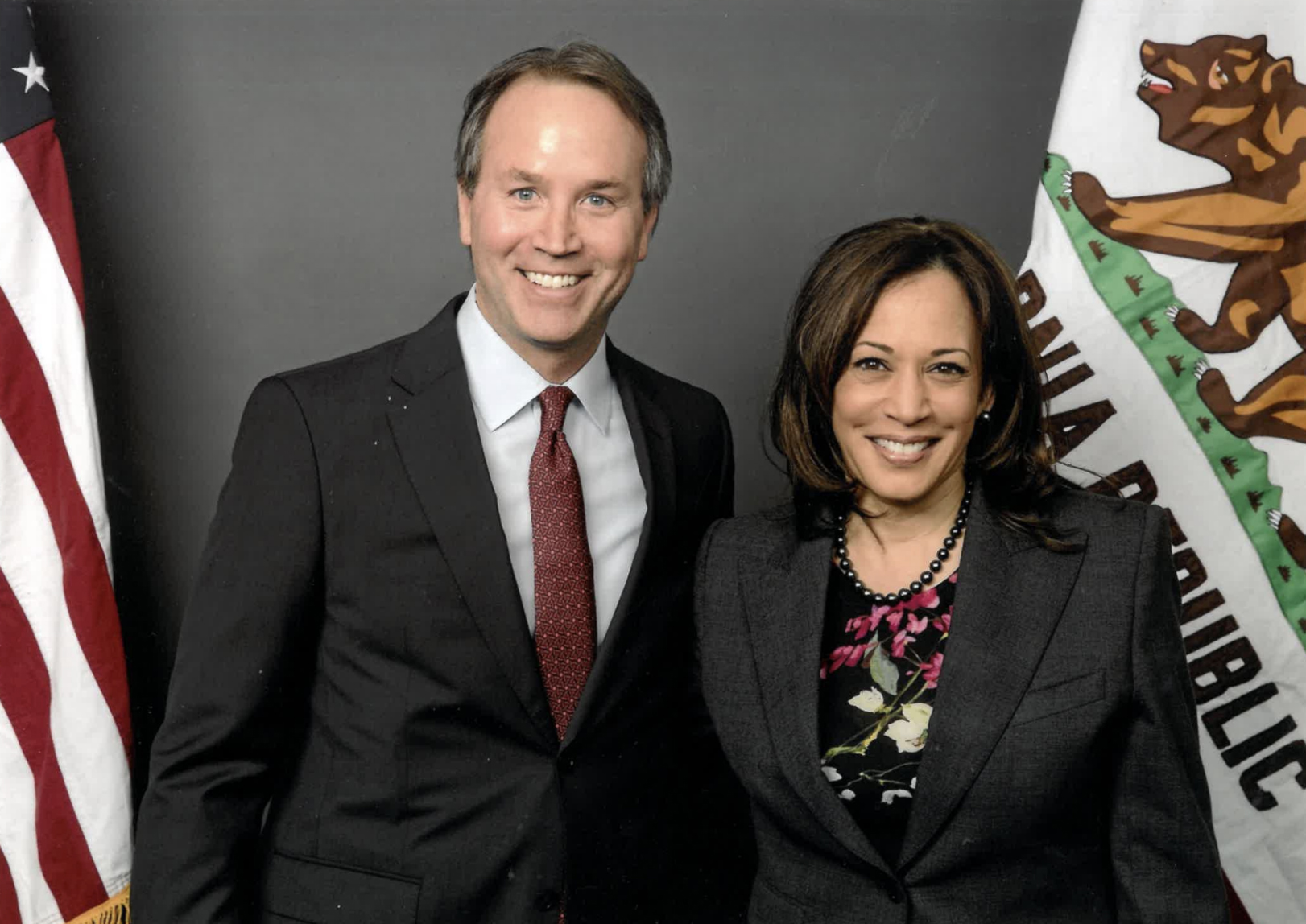 Two people in suits stand smiling before US and California flags.