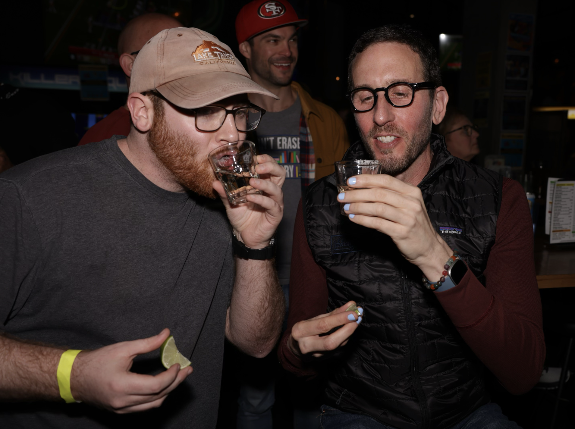 Two men are taking a shot at a bar; one is holding a lime wedge, both are wearing casual attire.