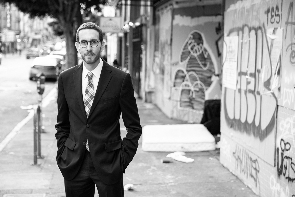 Sen. Scott Wiener in a suit stands on a graffiti-covered street, with a mattress and trash nearby. Black and white photo.