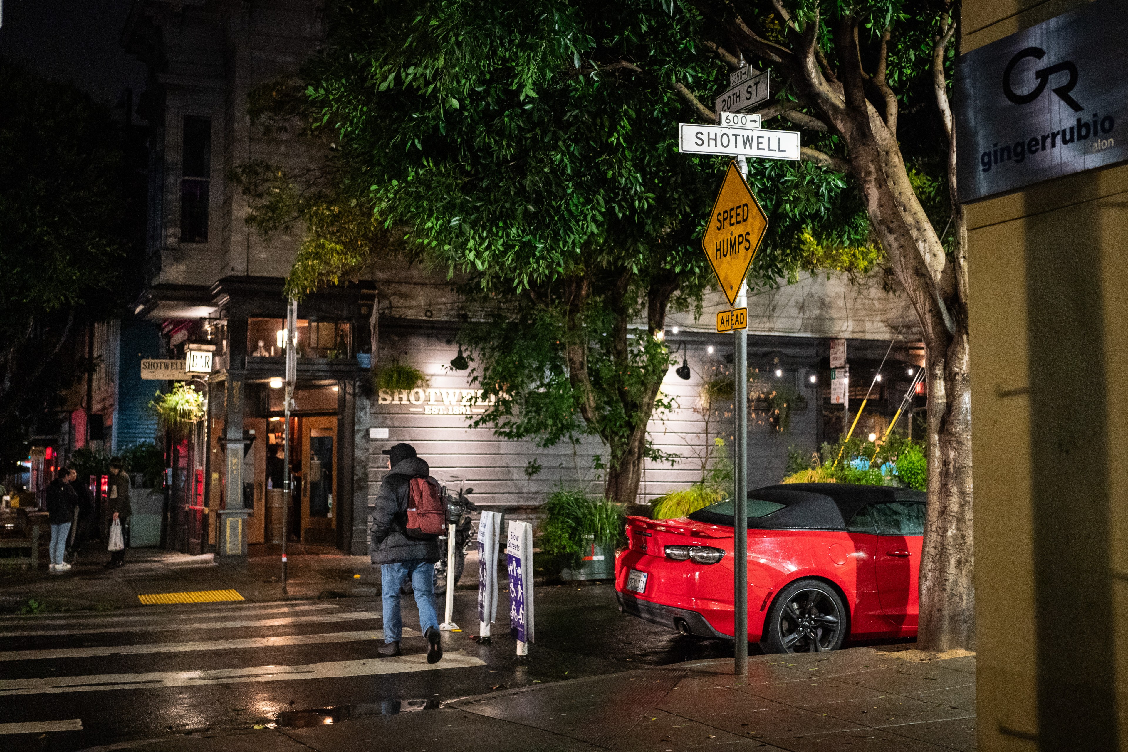 A rainy city corner at night with a pedestrian, a red car, and street signs, including one for Shotwell Street.