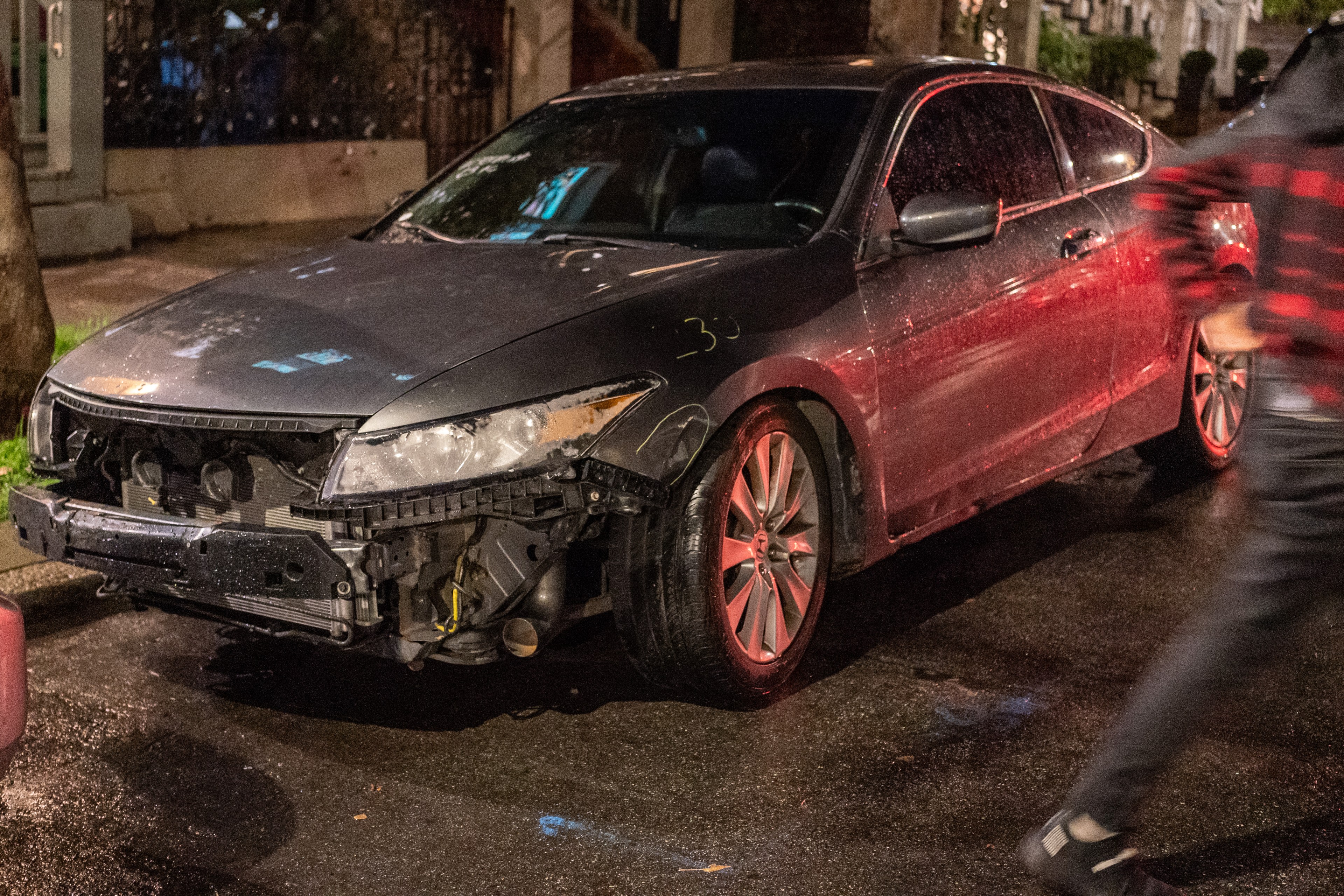 A damaged car with missing front bumper, exposed engine, and a blurred person walking by.