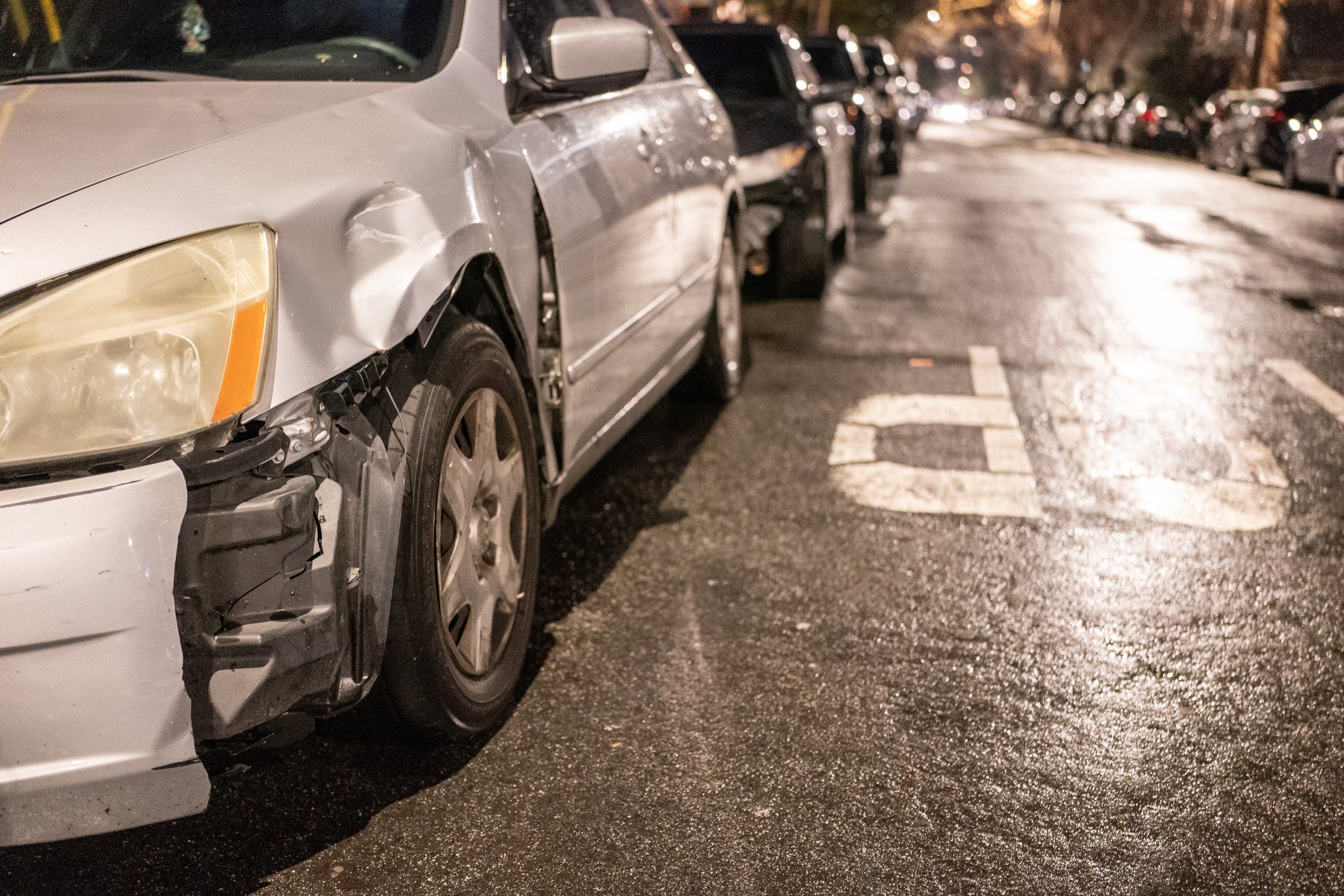 A white car with front-left damage, parked on a wet street at night, with other cars and street markings visible.