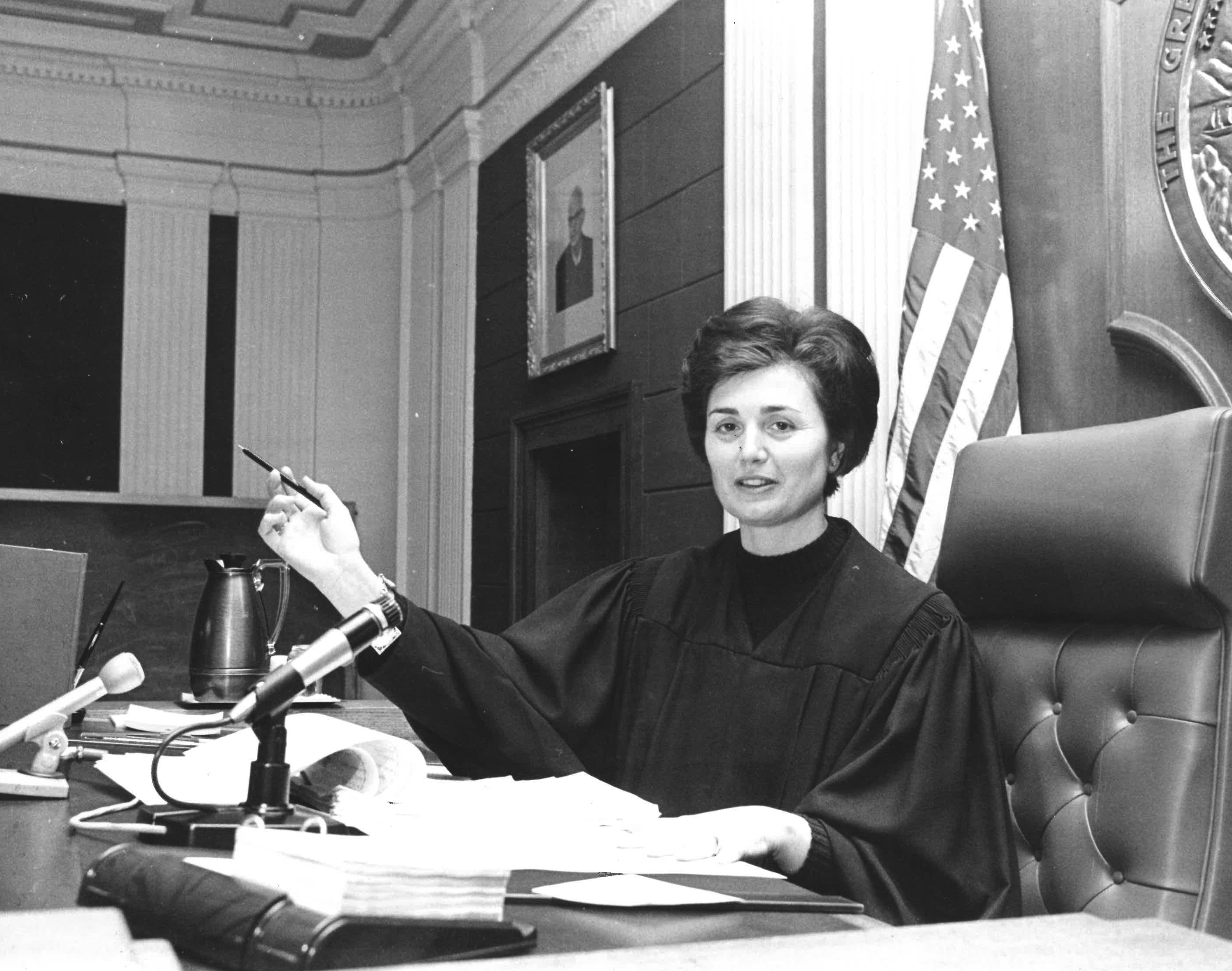 A woman in a judge's robe sits at a bench with a pen, papers, a microphone, and the U.S. flag behind her.