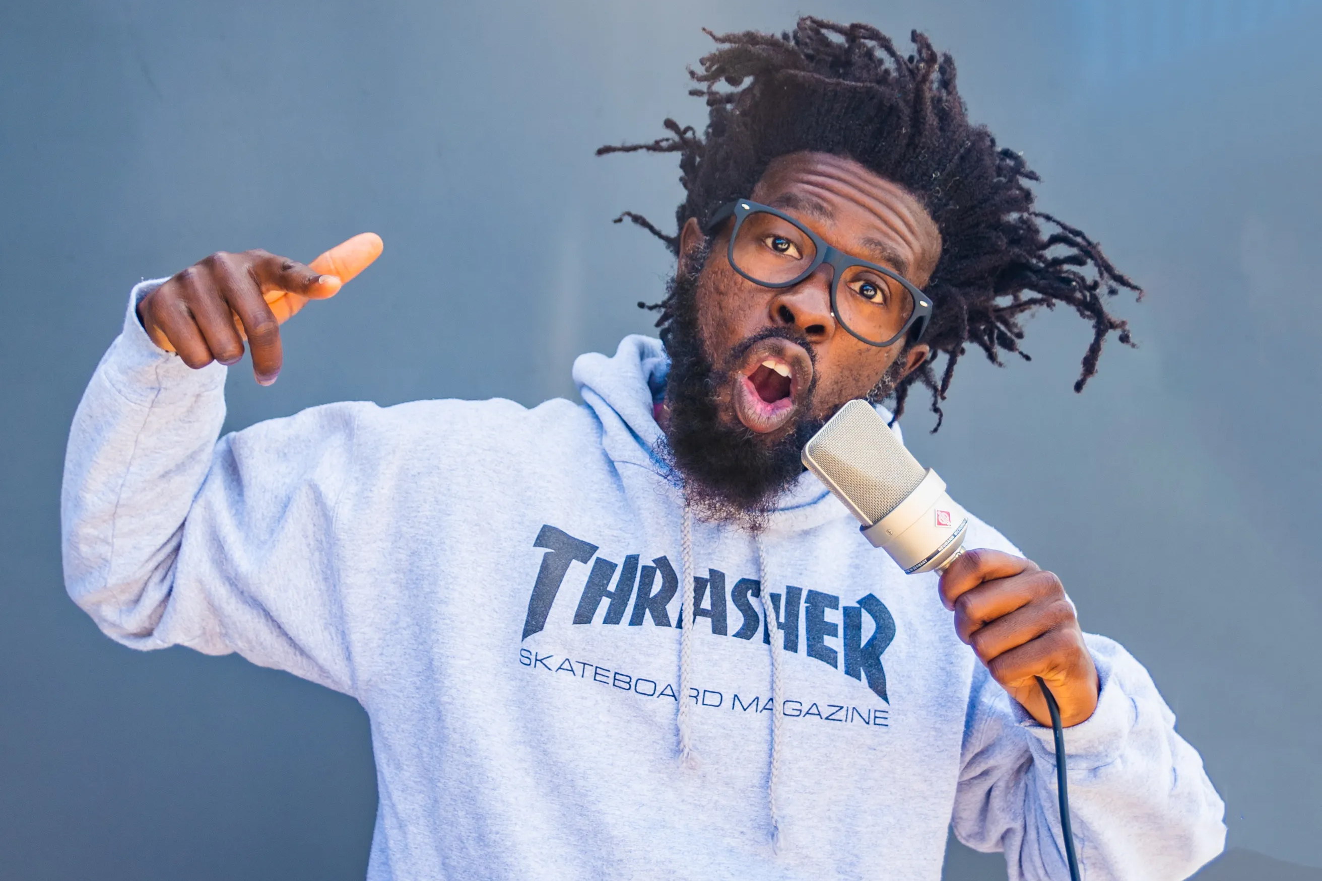 A man with dreadlocks, glasses, and a "Thrasher" hoodie holds a mic, pointing and singing with expression.