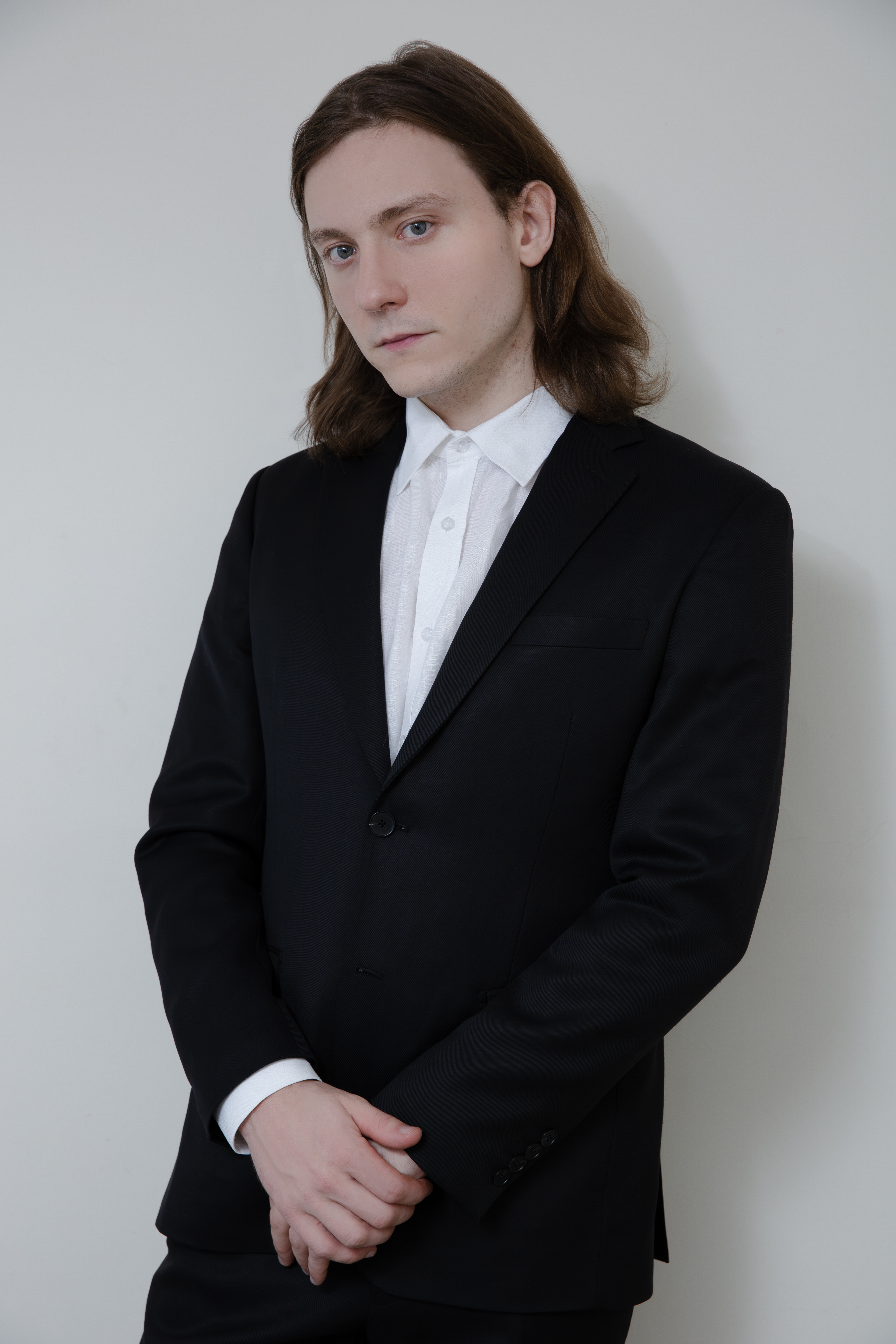 Man with long hair in a black suit and white shirt, hands clasped in front, against a white background.