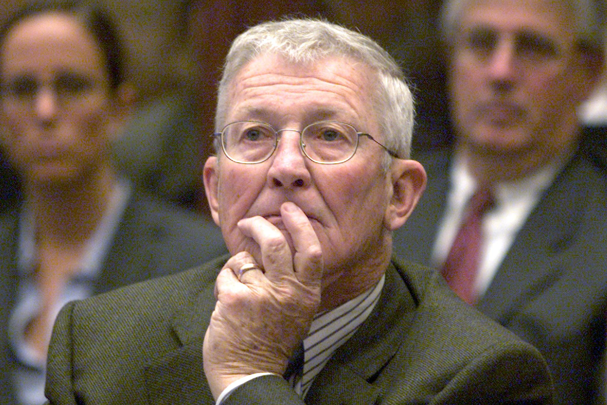 An older man with glasses, in a suit, thoughtfully rests his chin on his hand. Two blurred figures are behind him.