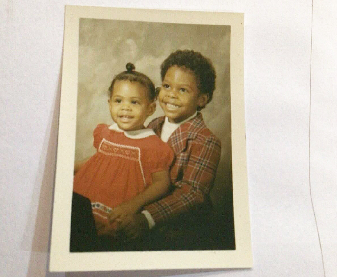 A vintage photo of two young children smiling; a girl in a red dress and a boy in a plaid shirt.