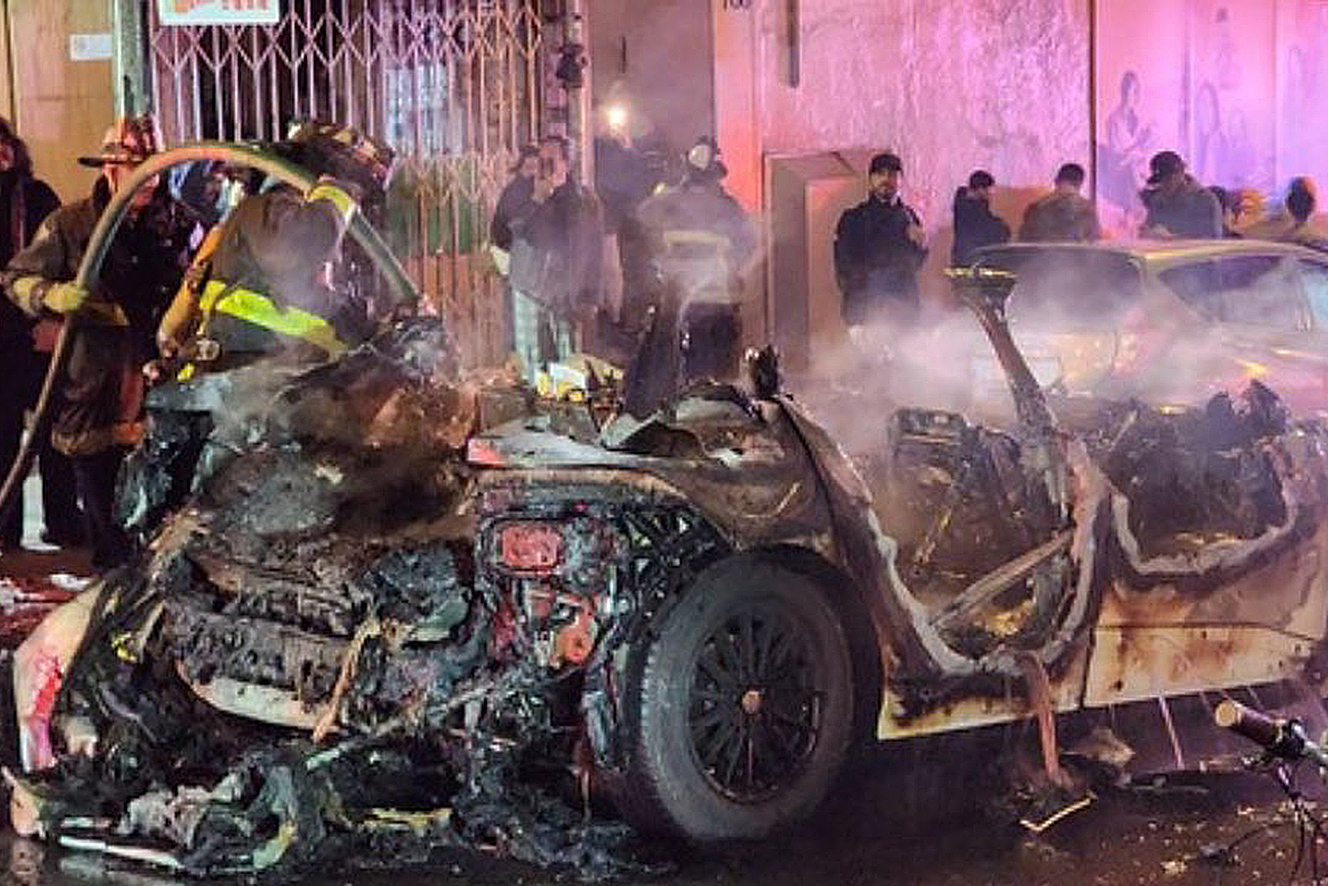 A firefighter hoses down a charred car wreck at night, with onlookers behind barriers.