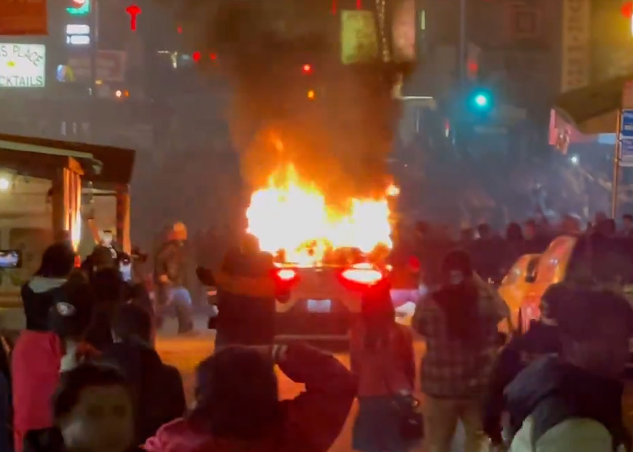 A car is engulfed in flames on a street at night, with onlookers capturing the scene on their phones.