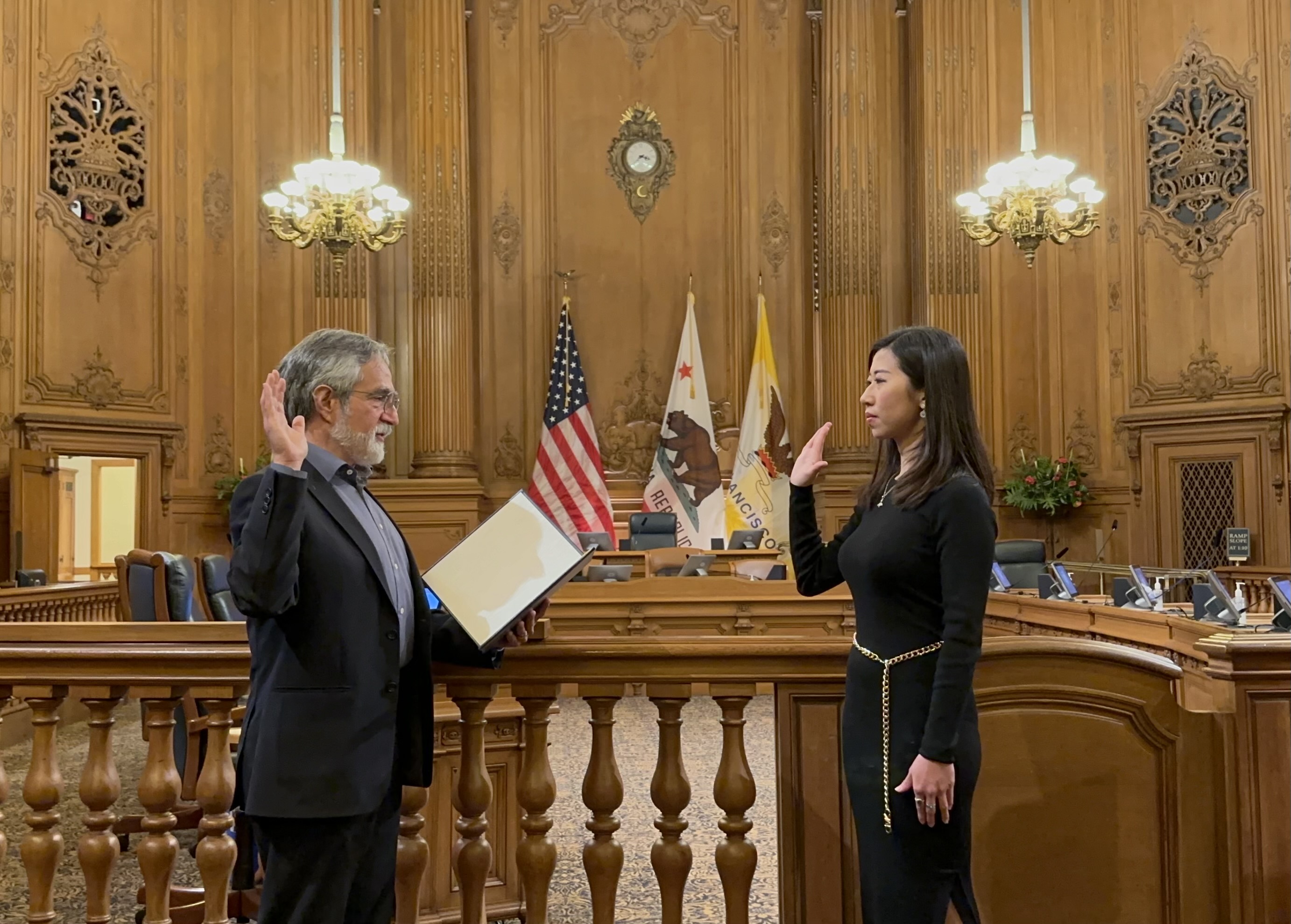 An oath-taking ceremony in a wood-paneled room with two people raising their right hands, an American flag, and ornate décor.