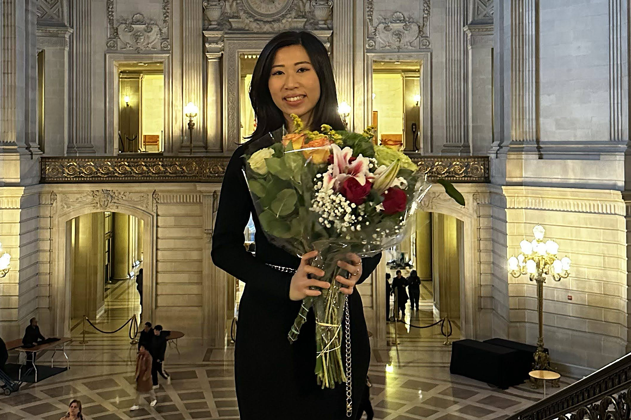 A woman in a black dress holds a bouquet of flowers, standing in an ornate hall with grand lighting.