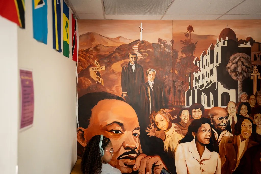 A mural depicts historical figures, with mountains, buildings, and a cross, viewed past a wall adorned with flags.
