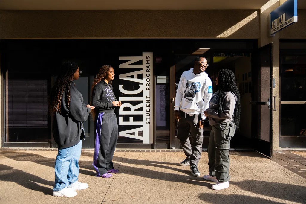 Four people conversing outside a building labeled &quot;AFRICAN STUDENT PROGRAMS.&quot; They wear casual attire and some smile in the sunlight.