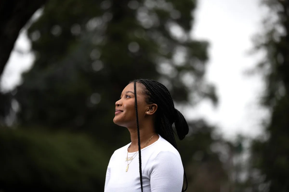 A woman with braided hair, wearing a white top, is looking upwards with a serene expression, trees softly blurred in the background.