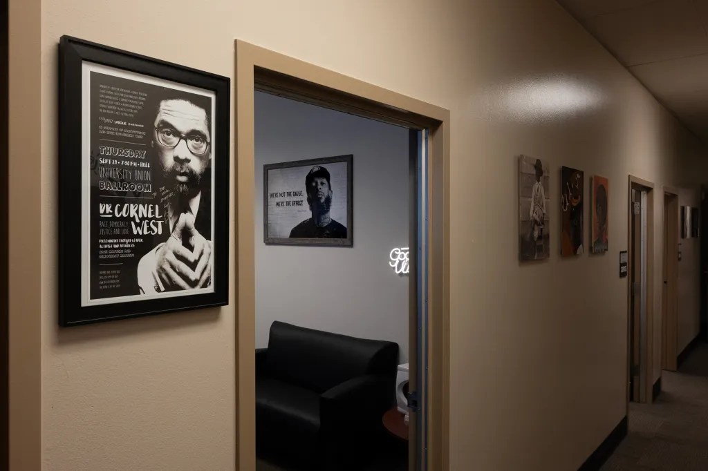 An office hallway with framed pictures, including a poster of a man with glasses and a beard, and a seating area with a black sofa.