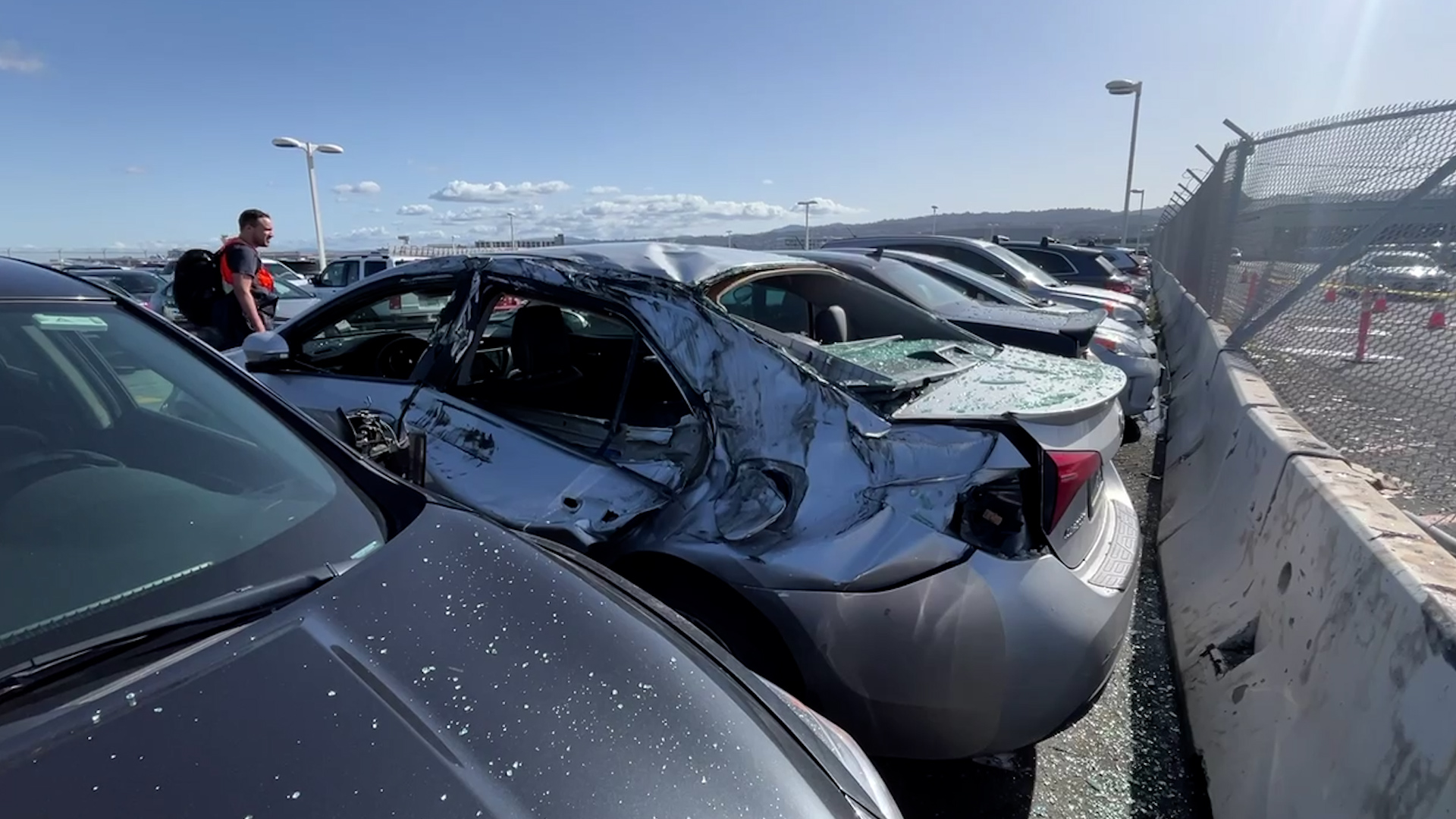 A damaged car in a parking lot, surrounded by intact vehicles, with a person nearby.