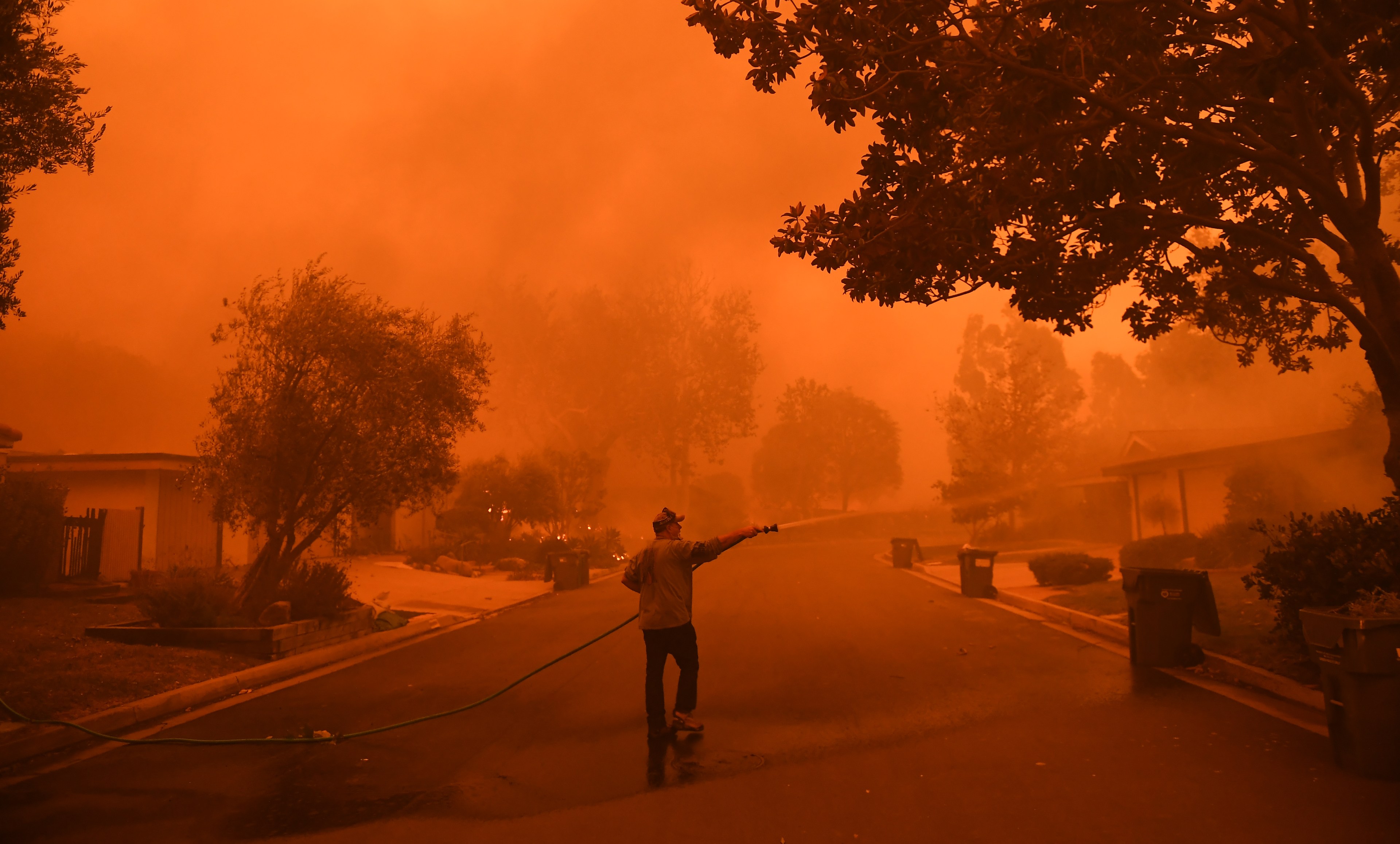 A person hoses down a street under a sky glowing orange due to wildfire smoke.