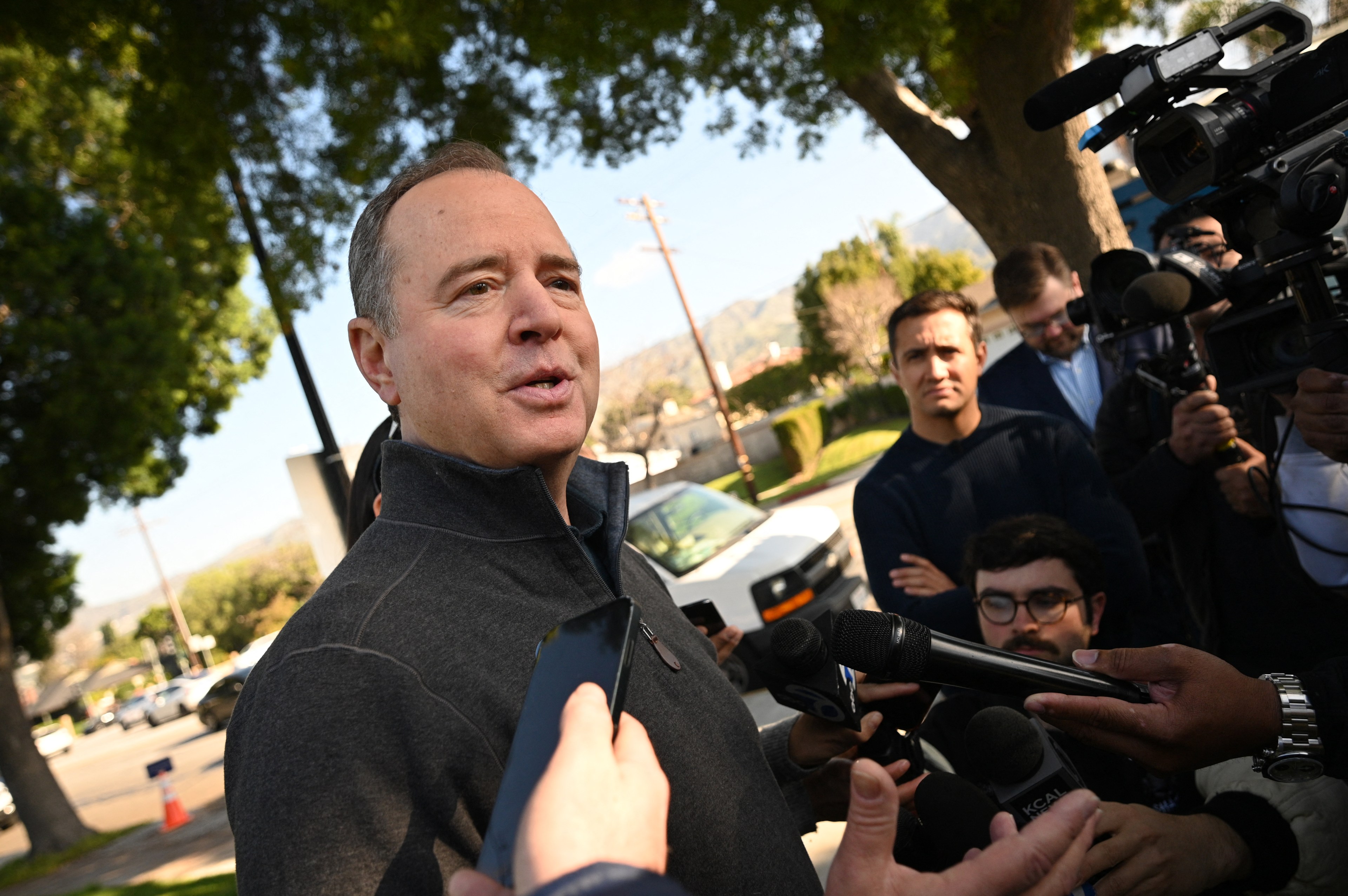 A man speaks to reporters holding microphones, with cameras and people behind him.