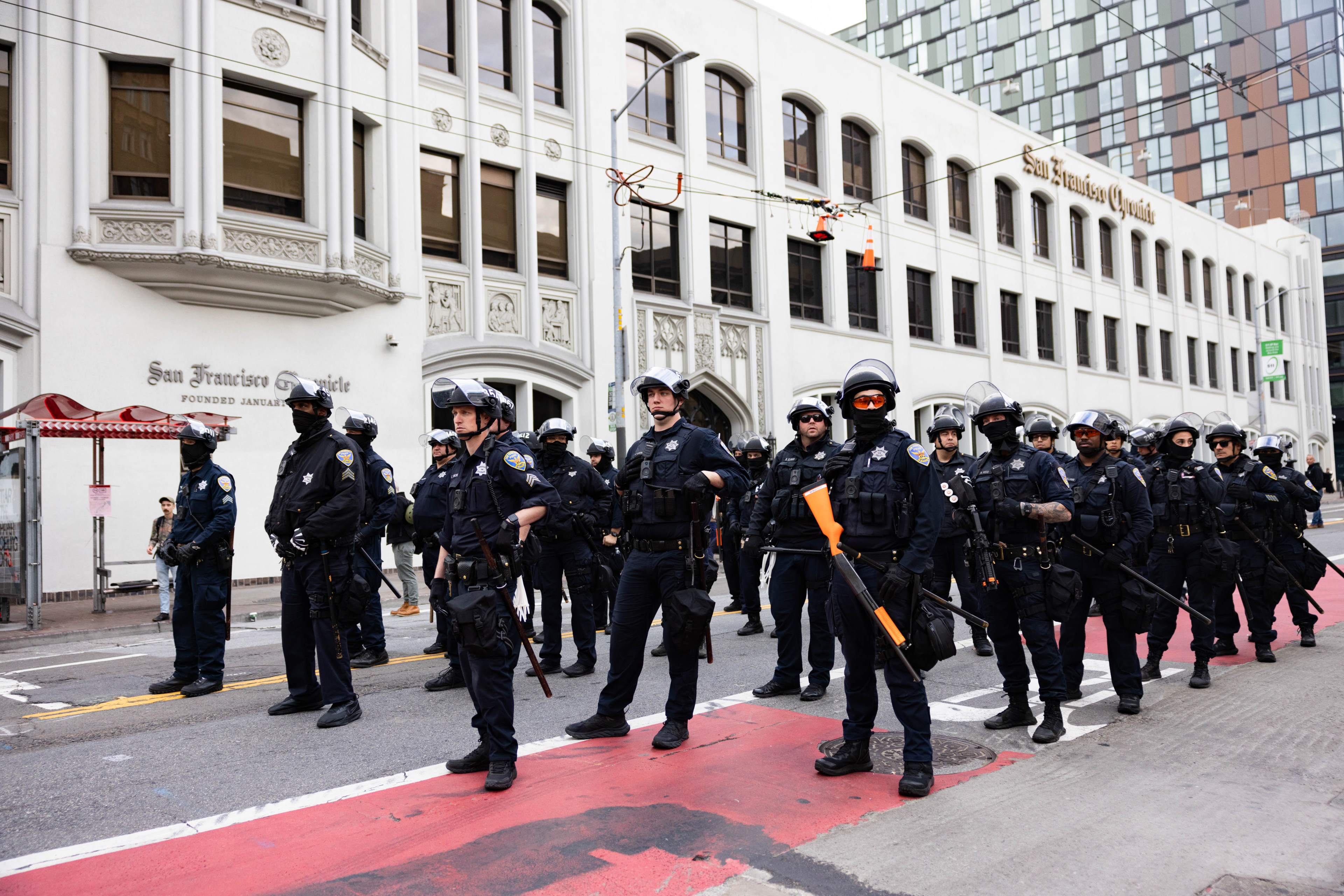 A group of riot police in gear stands ready on a city street, with a building labeled &quot;San Francisco Chronicle&quot; behind them.