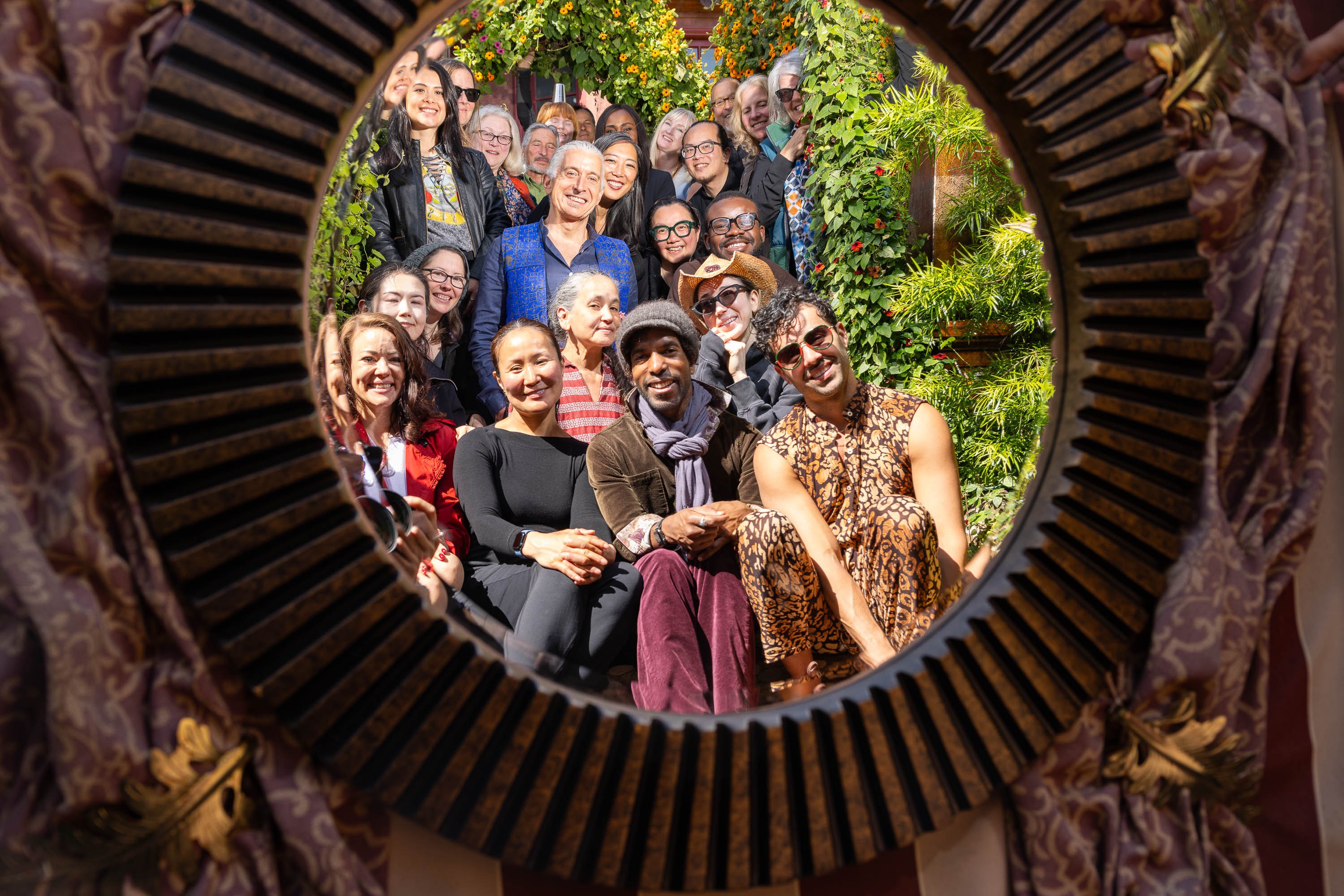 Group of smiling people seen through an ornate circular frame, surrounded by lush greenery.