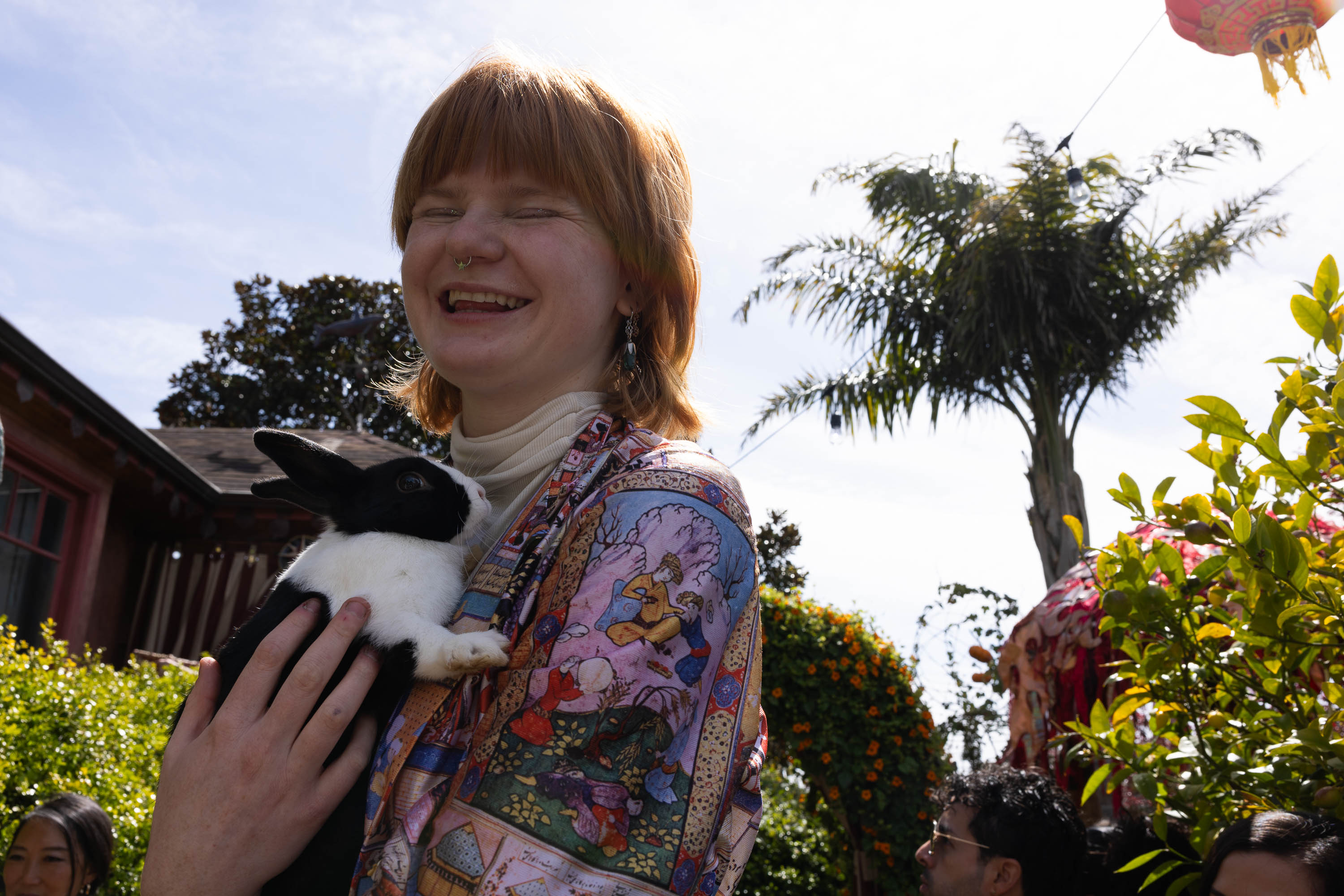 A person with a warm smile holding a black and white rabbit outdoors in sunlight.
