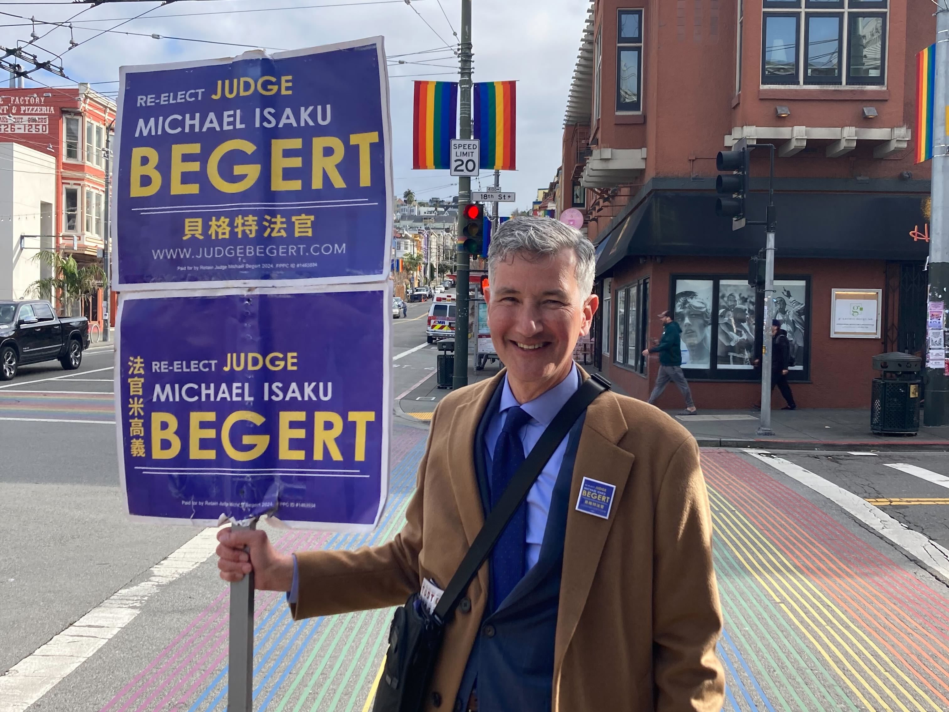 A smiling man on a colorful crosswalk holds a campaign sign for Judge Michael Begert.