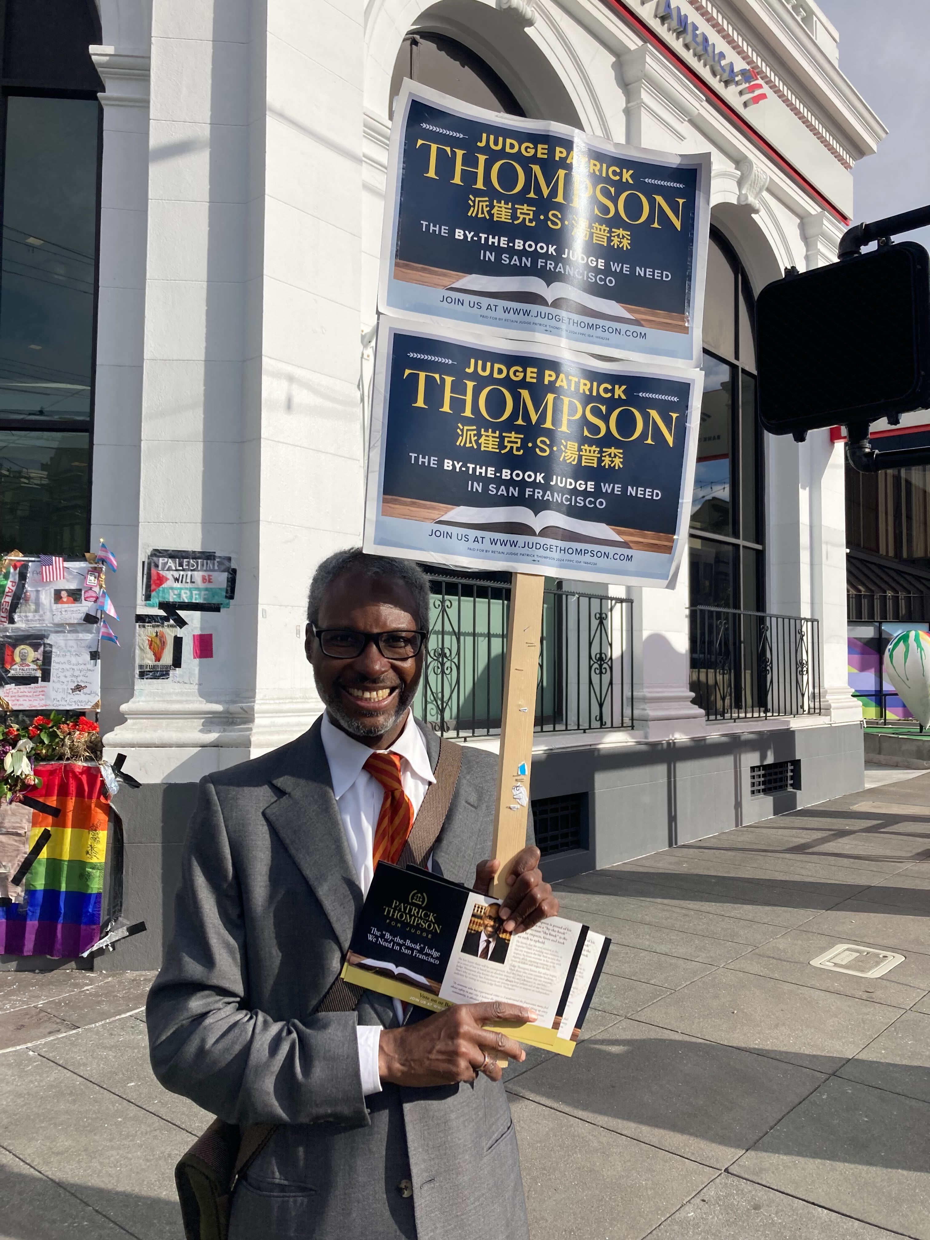 A man is smiling, holding campaign signs and a pamphlet promoting a judicial candidate. There's a pride flag in the background.