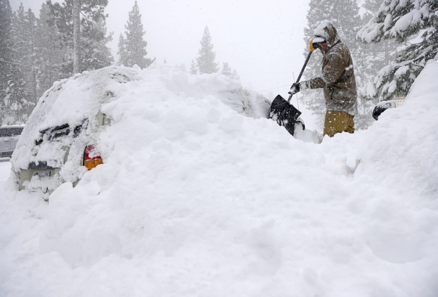 A person in winter gear shovels heavy snow engulfing a vehicle amidst a snowfall.