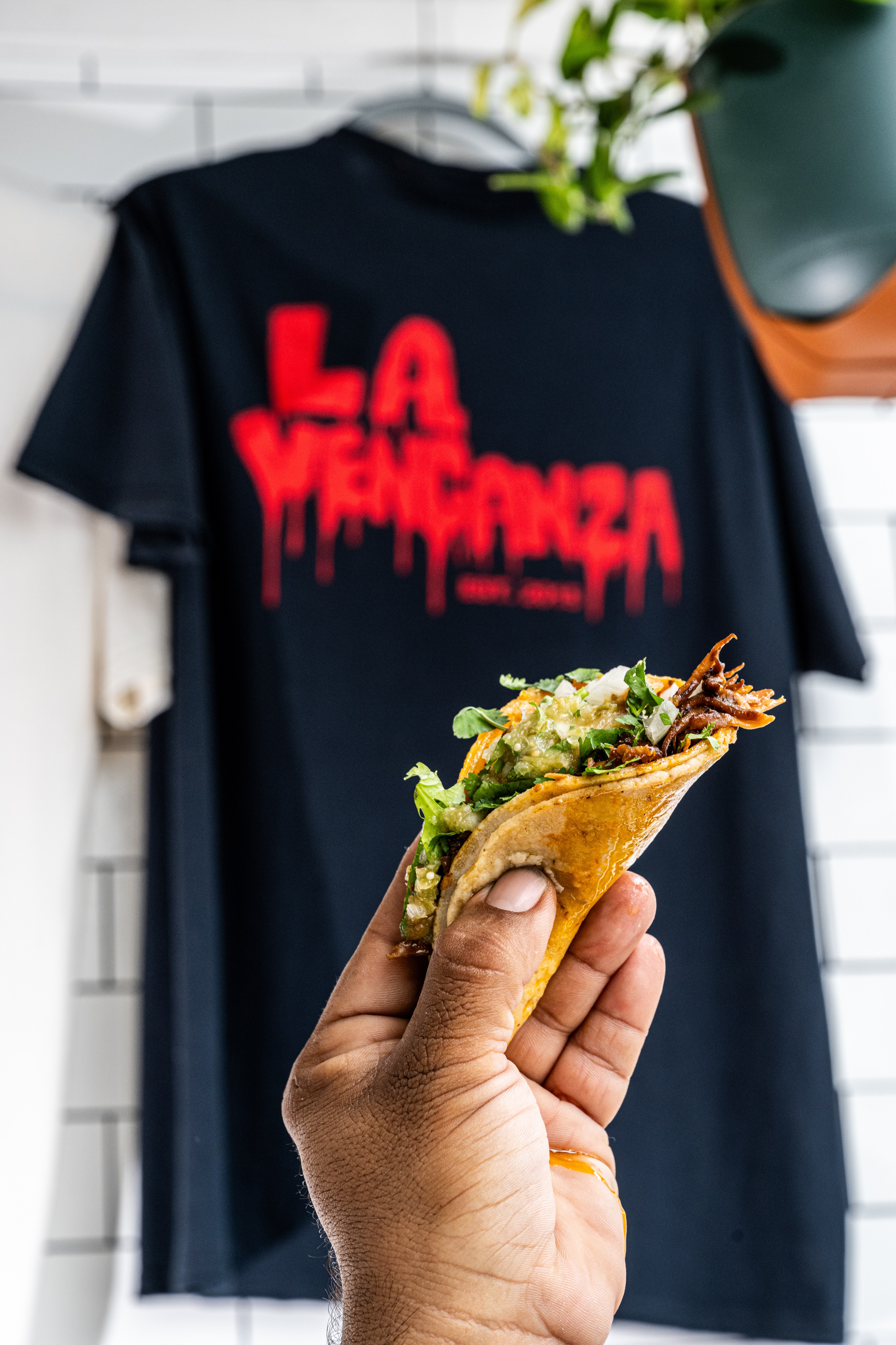 A hand holds a taco in front of a black T-shirt with &quot;LA VENGANZA&quot; in red text hanging on a wall.