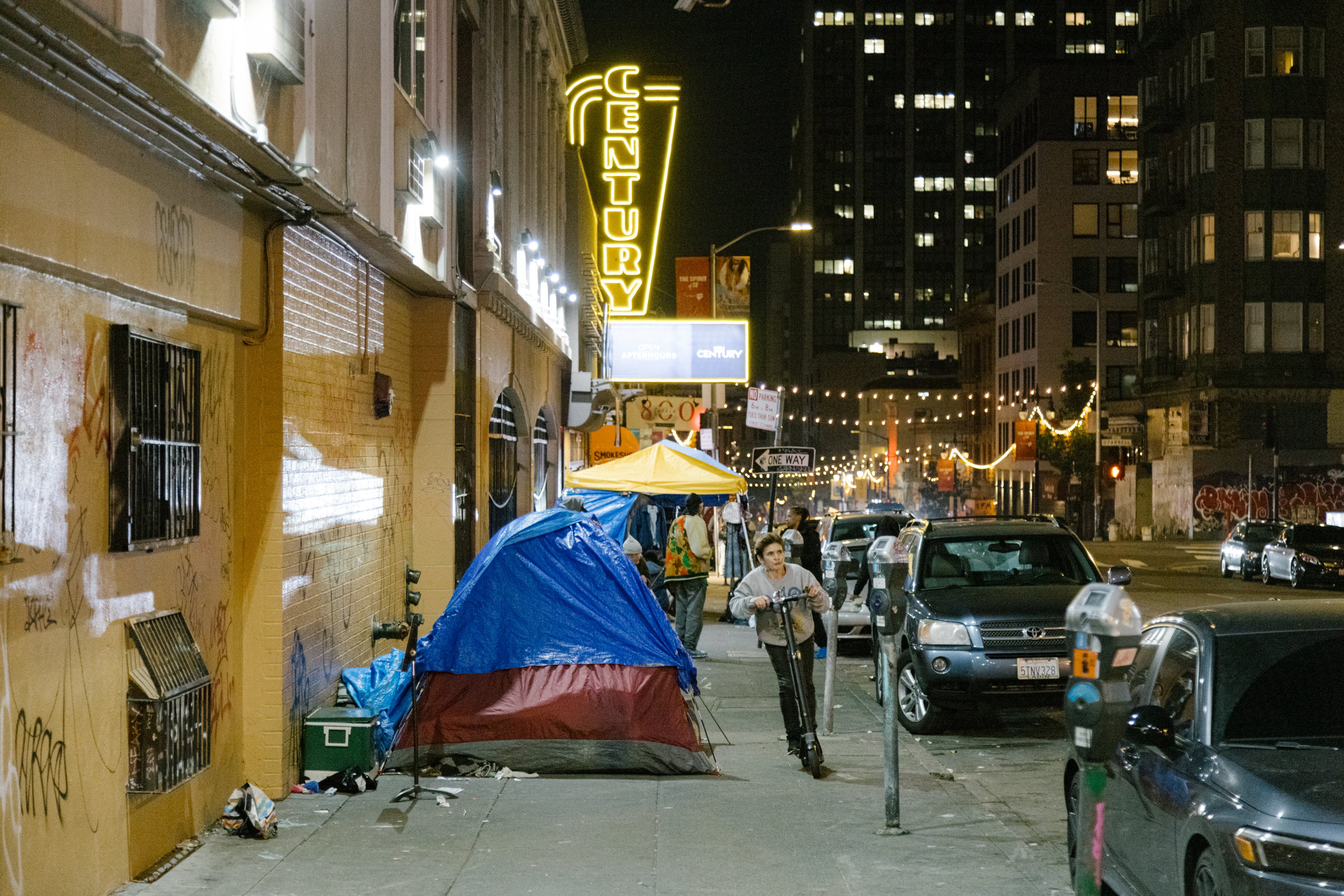 A city street at night with a blue tent on the sidewalk, neon signs, a parked scooter, and cars.