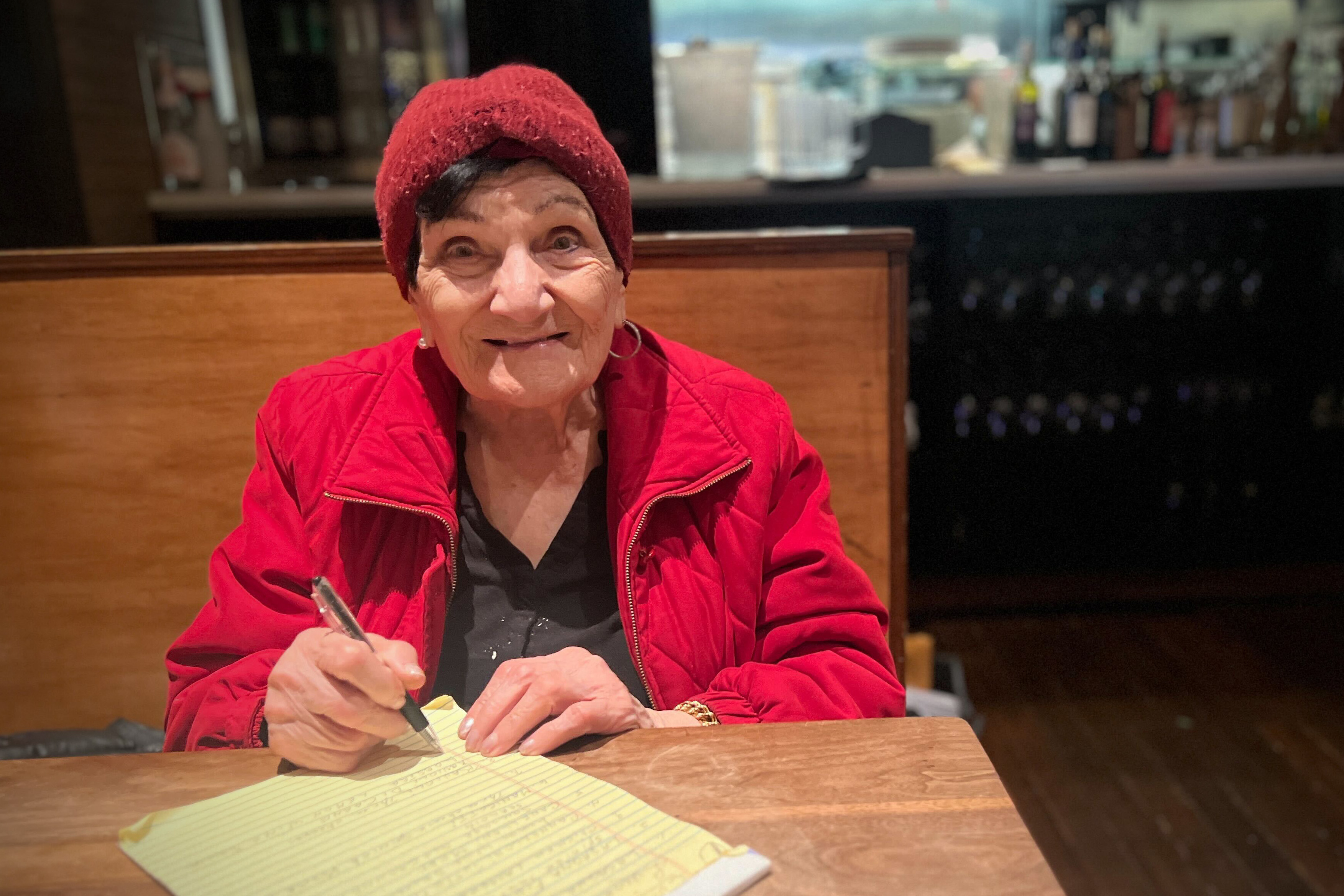 An elderly lady in a red hat and jacket smiles while writing on a yellow paper at a table.