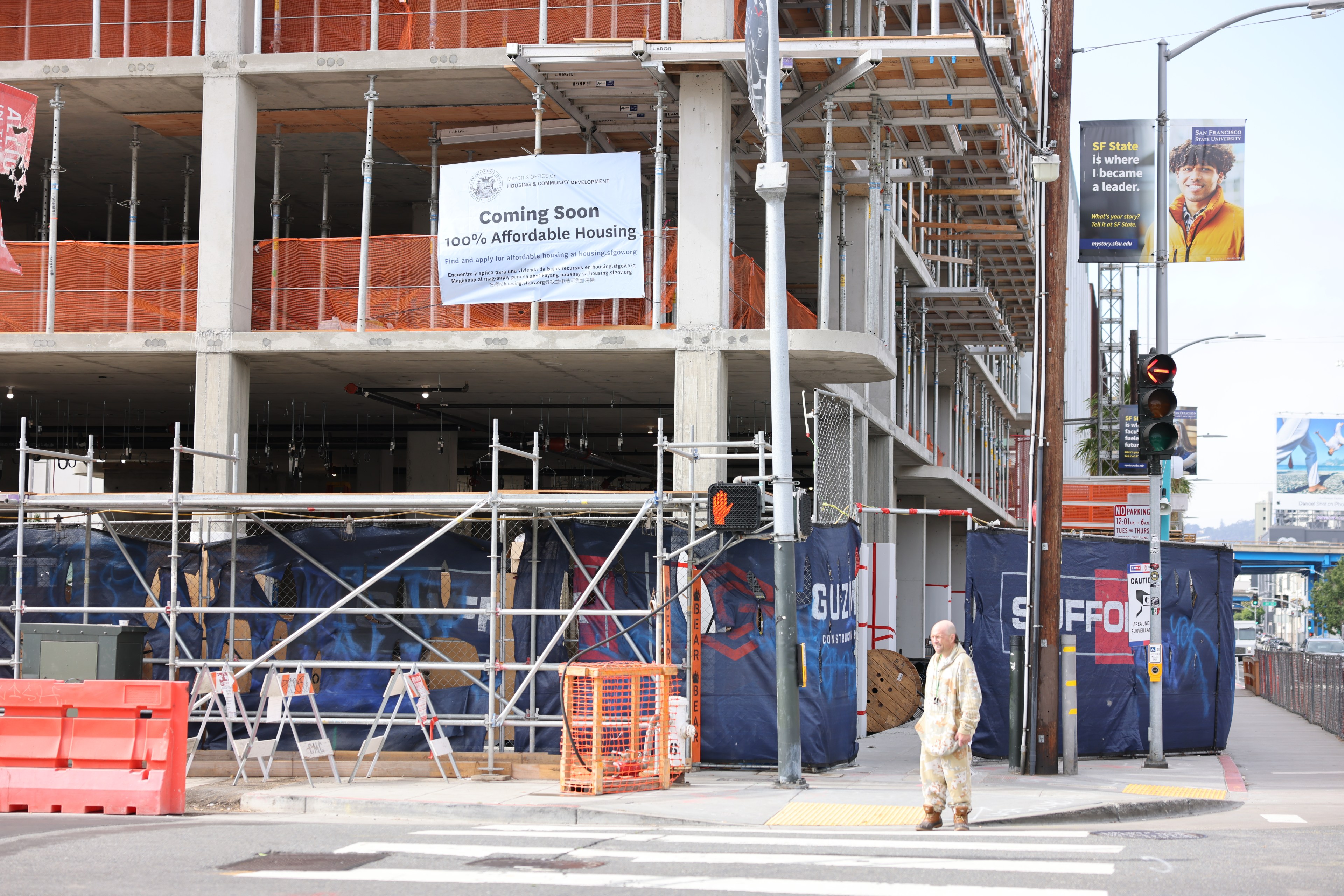 A construction site with scaffolding shows a &quot;Coming Soon 100% Affordable Housing&quot; sign. A man in paint-stained clothes stands at a crosswalk.