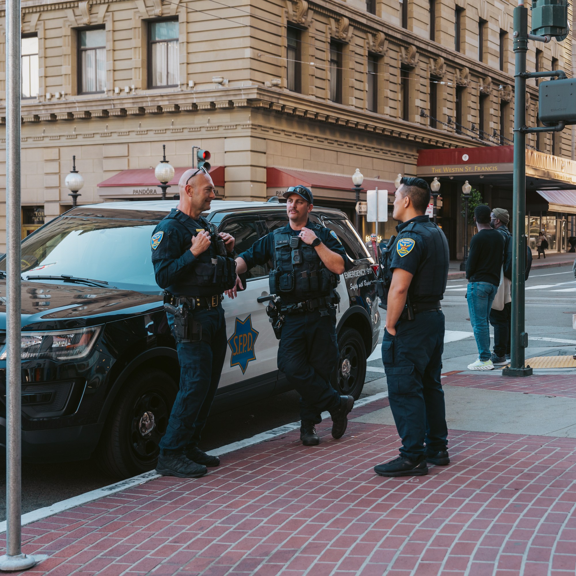 Three police officers stand talking beside a patrol car in a city street. Nearby, pedestrians, including a child, observe, and a cyclist passes by.