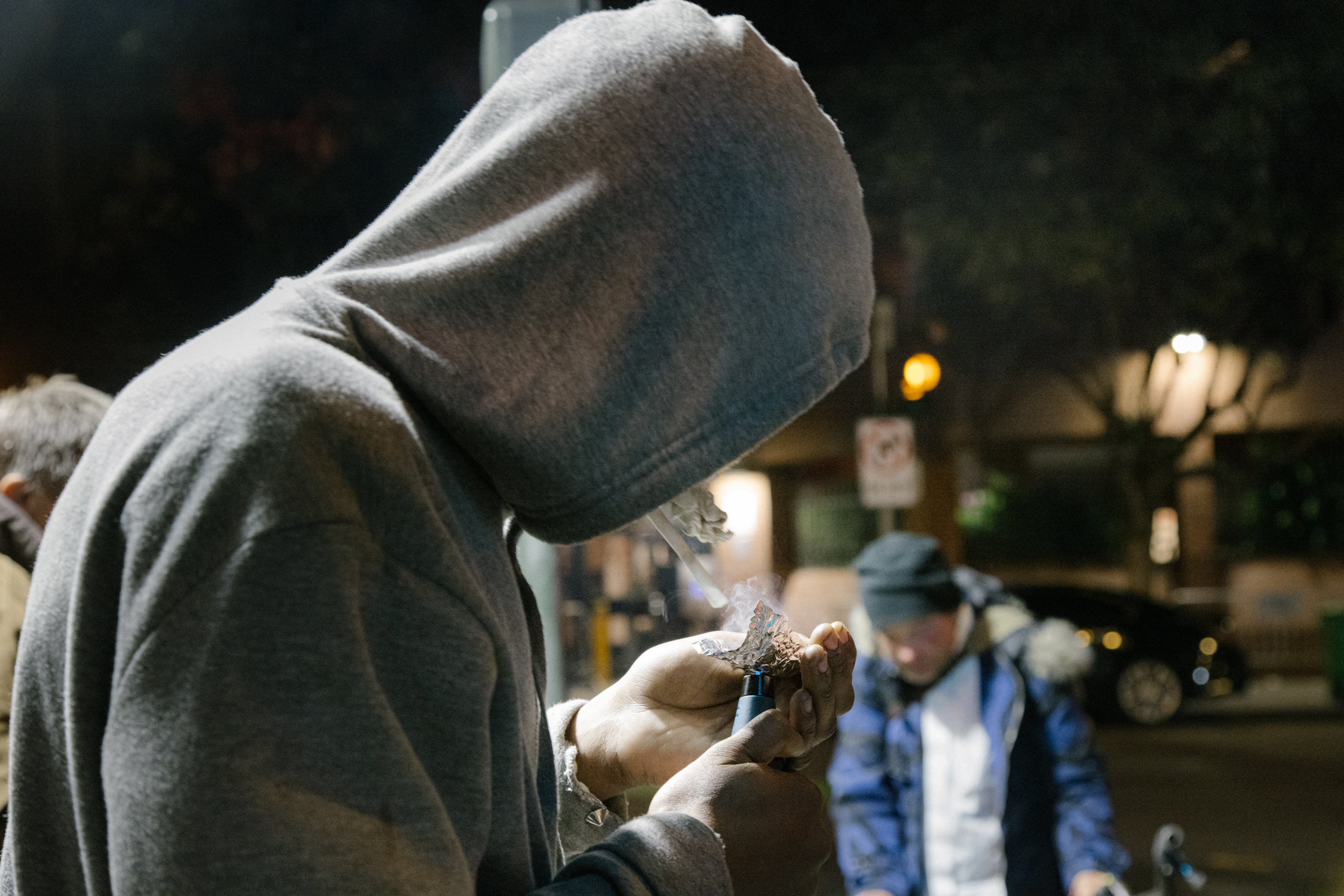 A person in a hood lighting a pipe, another blurred figure in background, nighttime urban setting.