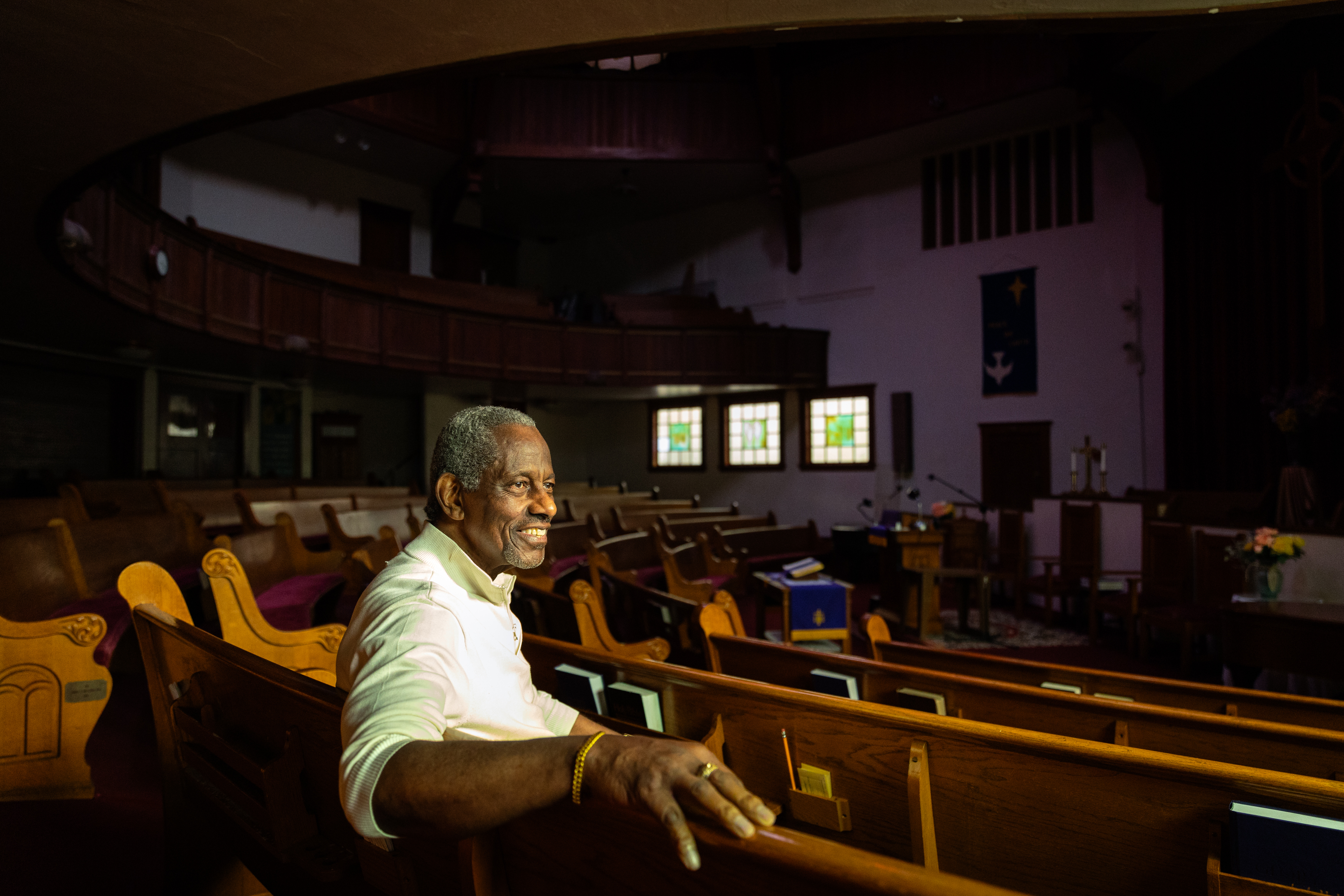 A smiling man sits in a sunlit church, amidst wooden pews and stained-glass windows.