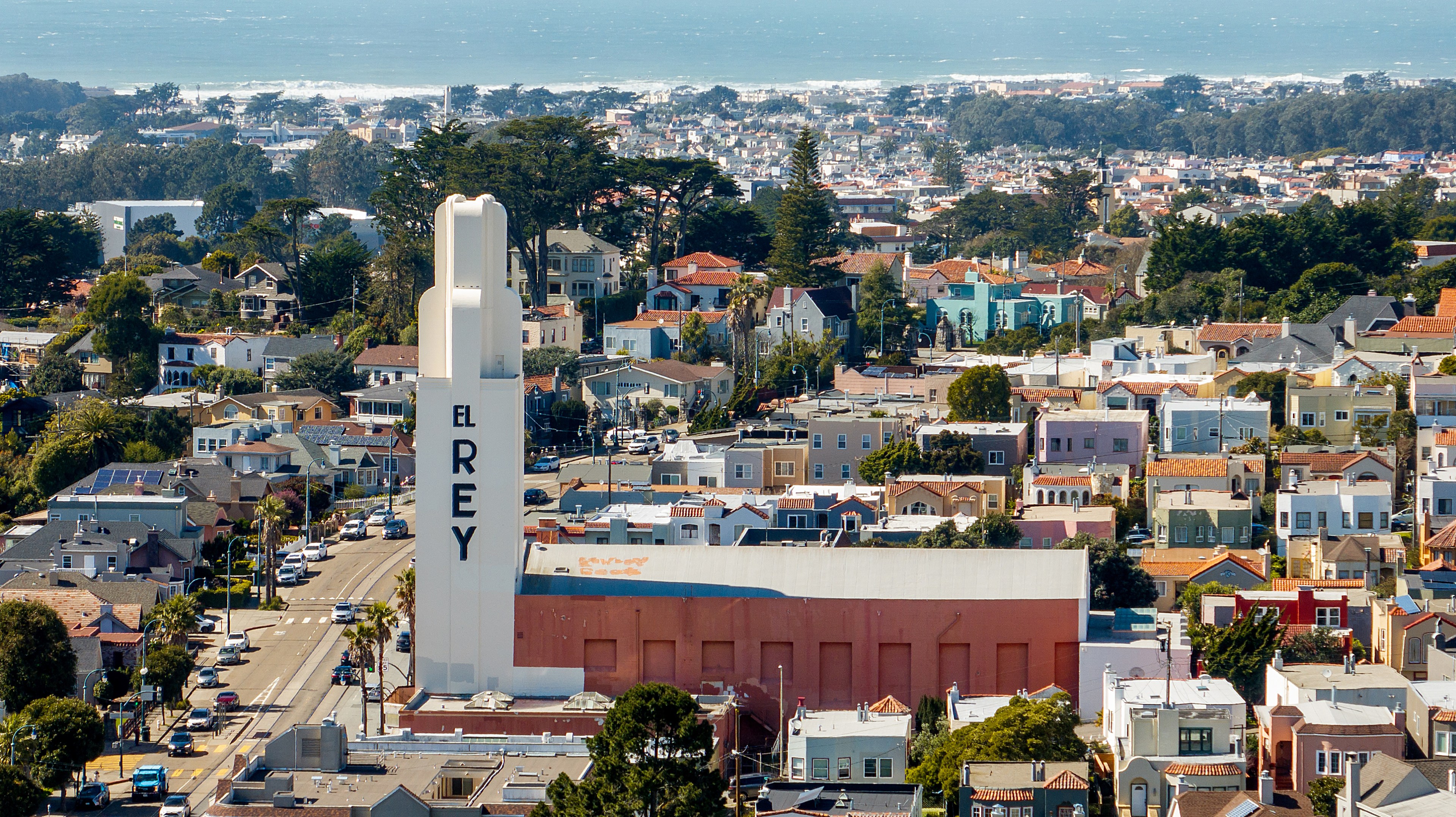 A large vertical sign reading "EL REY" atop a building, with rows of colorful houses and trees extending towards a hazy coastline in the background.