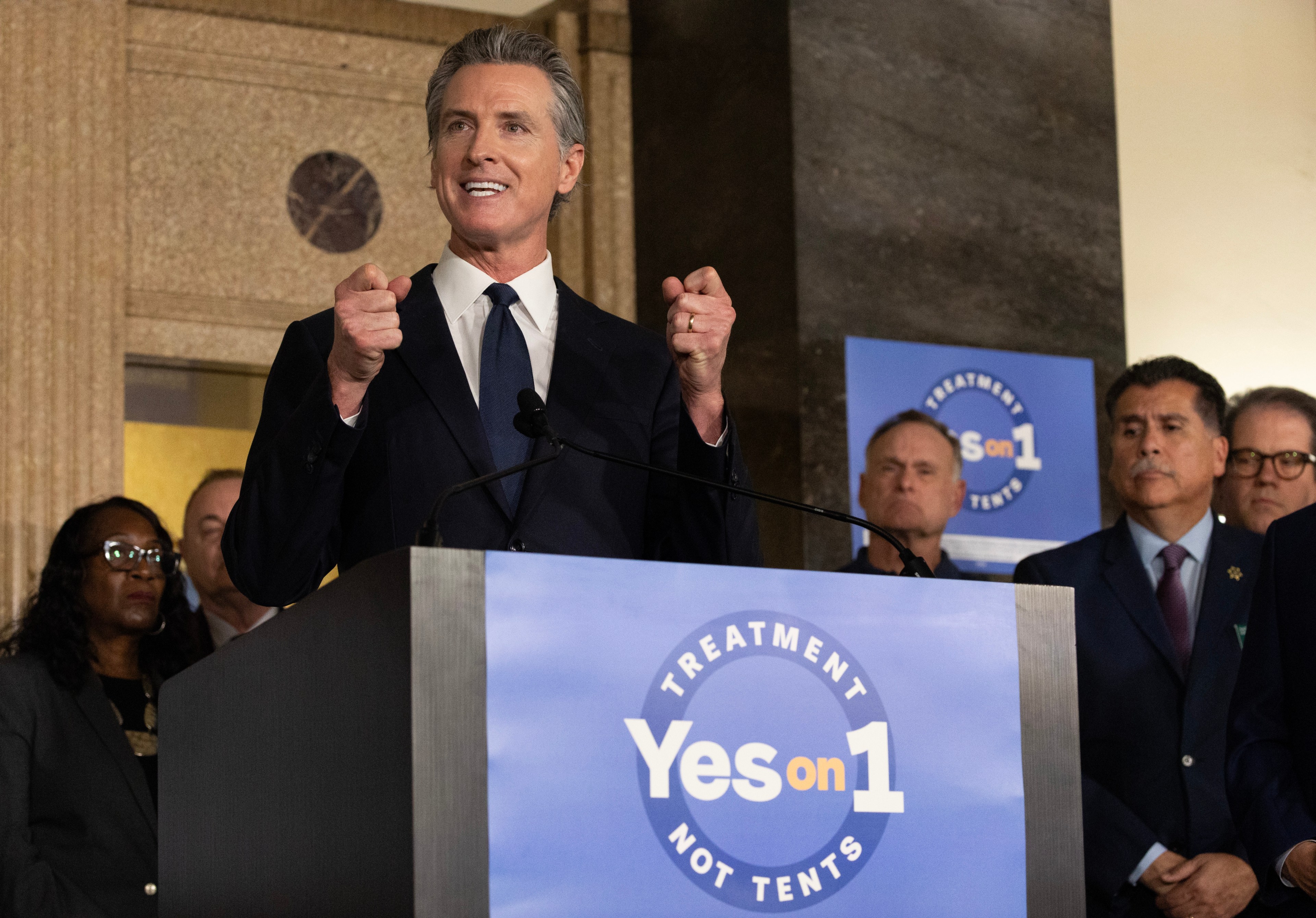 A man at a podium gestures passionately, with a "Yes on 1" sign, and attentive people around him.