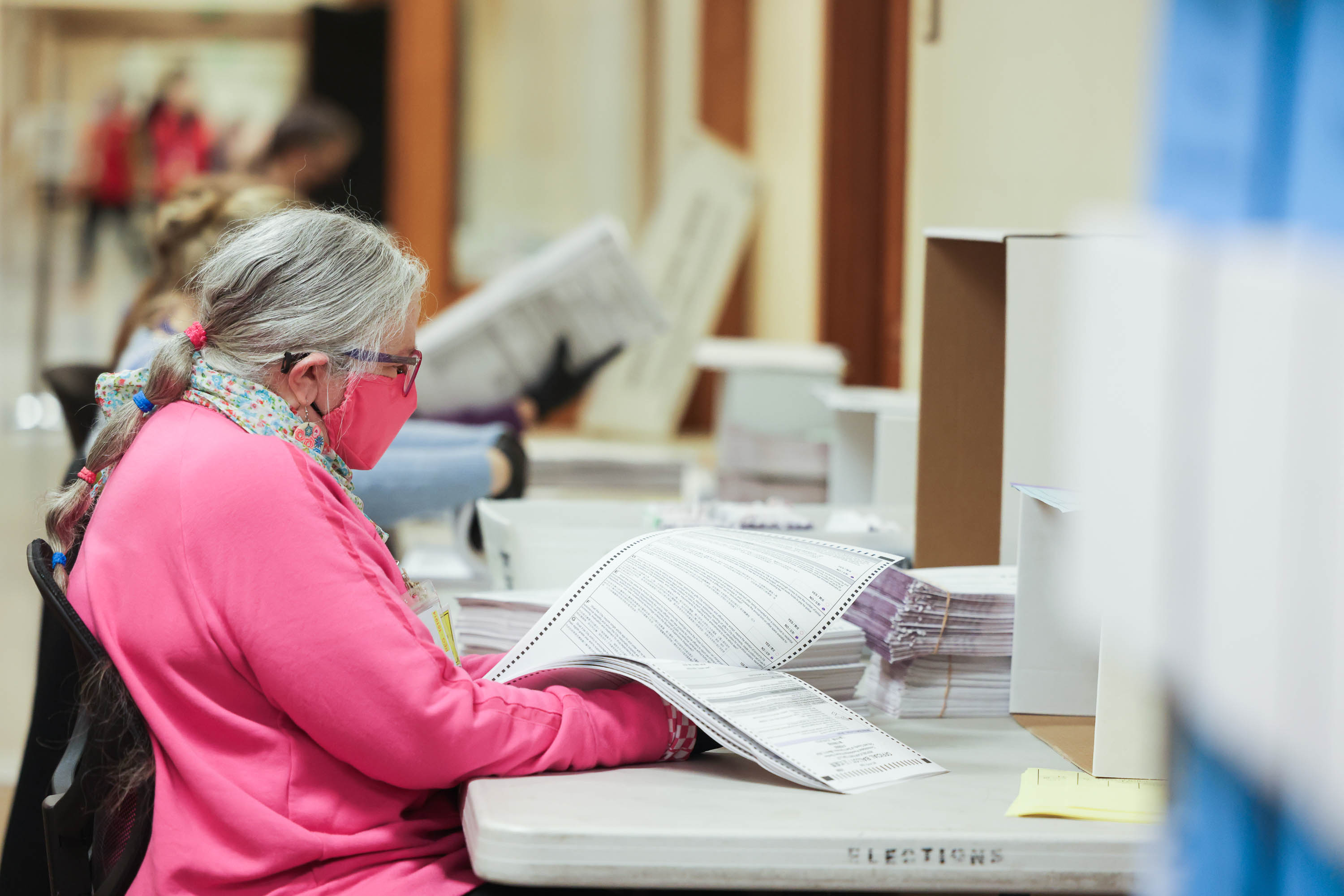 A person in a pink outfit and a face mask is examining papers in a room with labeled &quot;Elections&quot; bins.