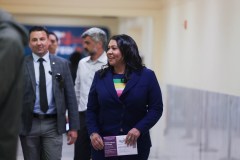 A woman in a blue blazer and rainbow shirt walks confidently, holding papers, followed by two men in suits.