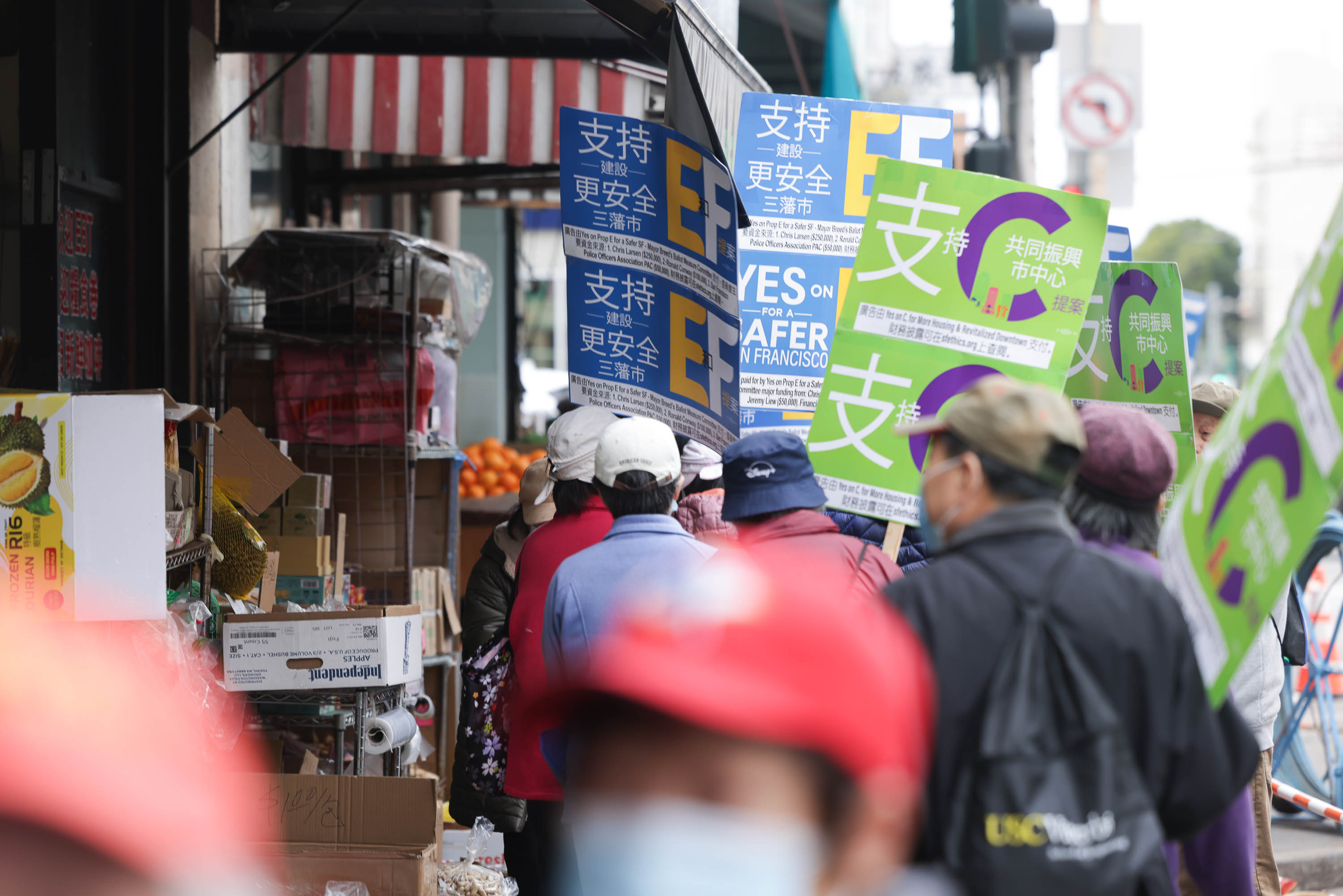 A bustling street scene in San Fran Francisco's Chinatown with people holding colorful protest signs with various text, including: &quot;YES ON E FOR A SAFER SAN FRANCISCO.&quot;